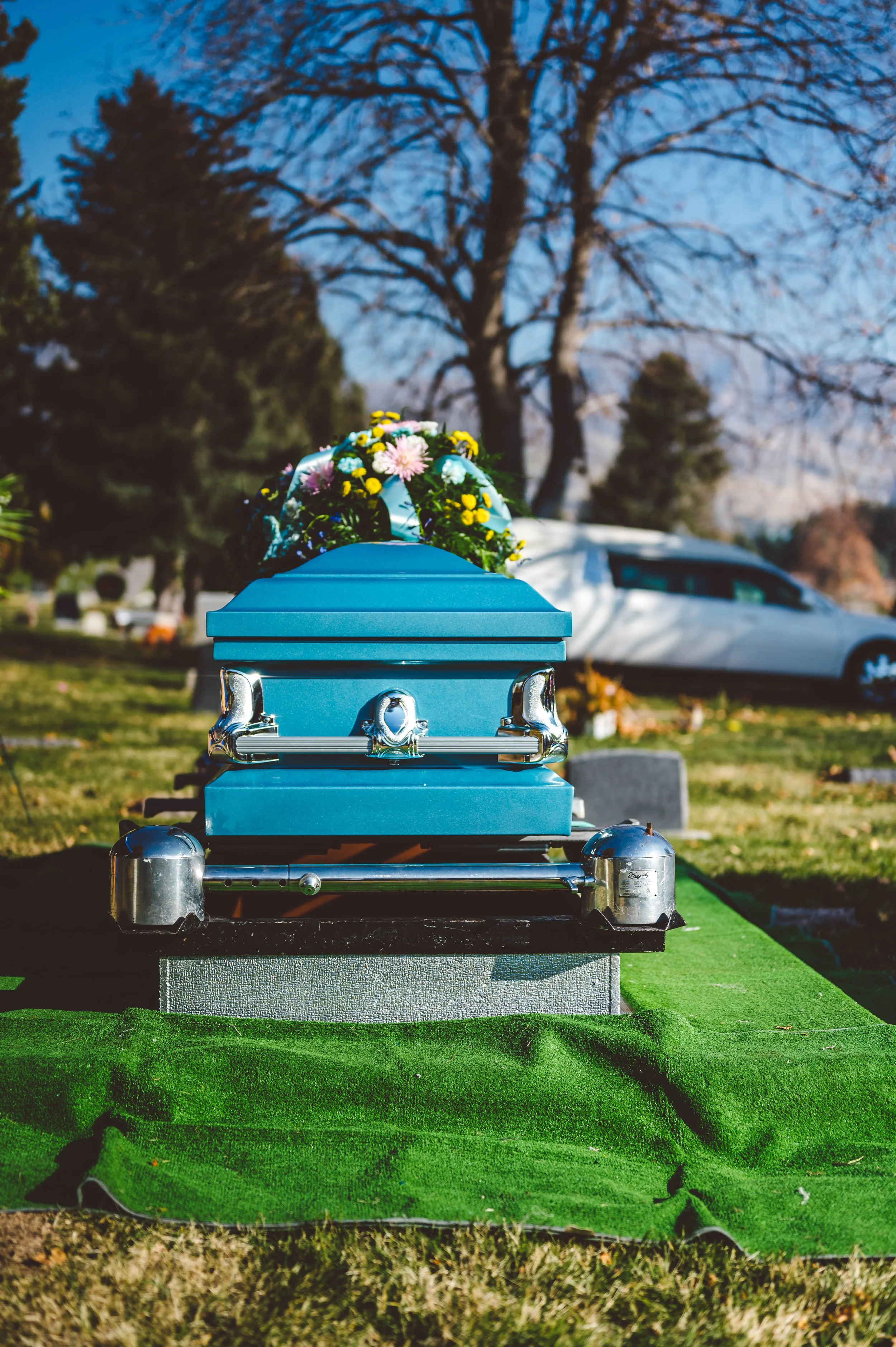 A blue casket with a floral wreath on top, placed on a green mat in a graveyard during daytime.