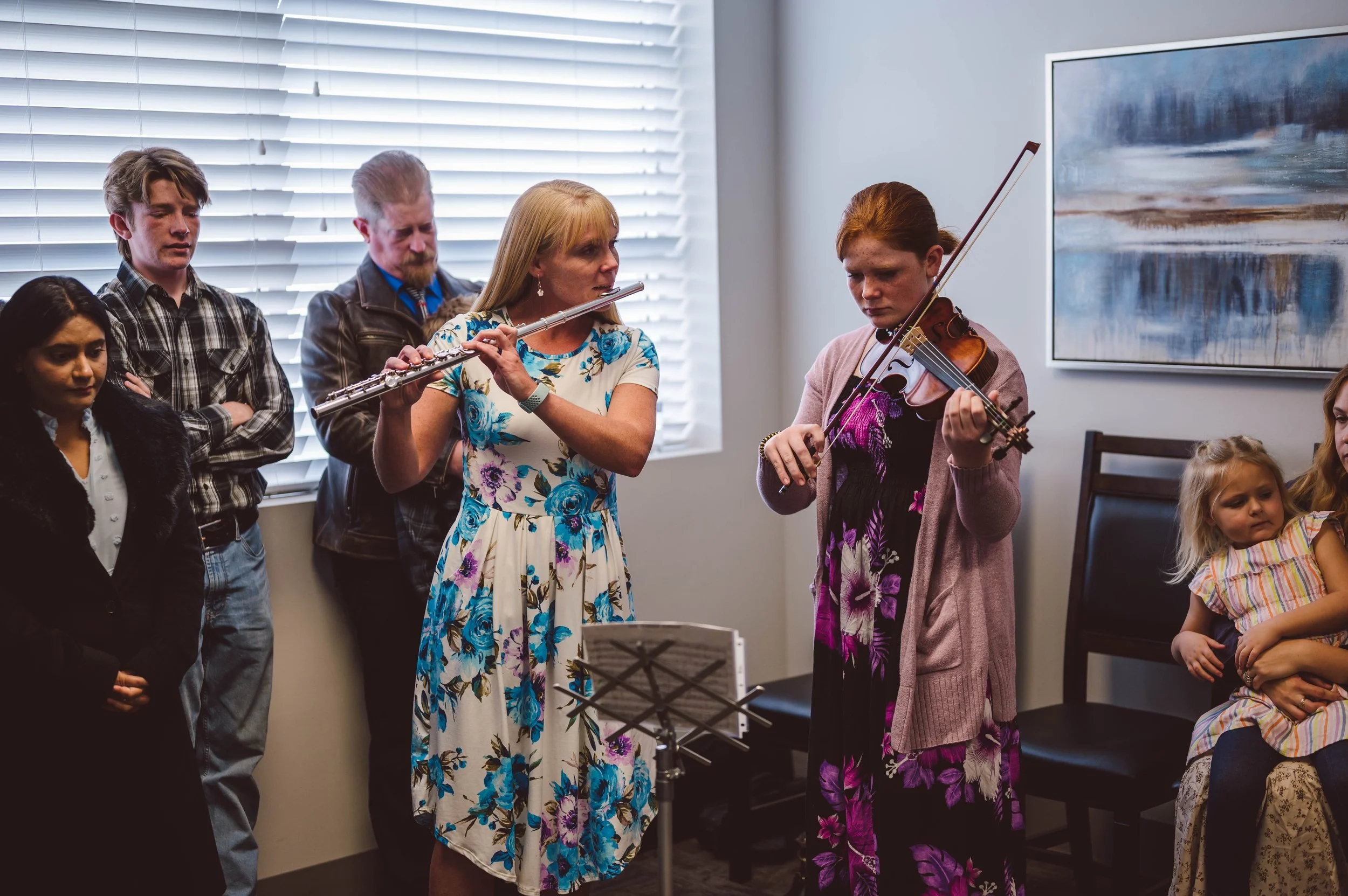 Two women are playing violins while others watch during a gathering in a room with a large window and abstract wall art.