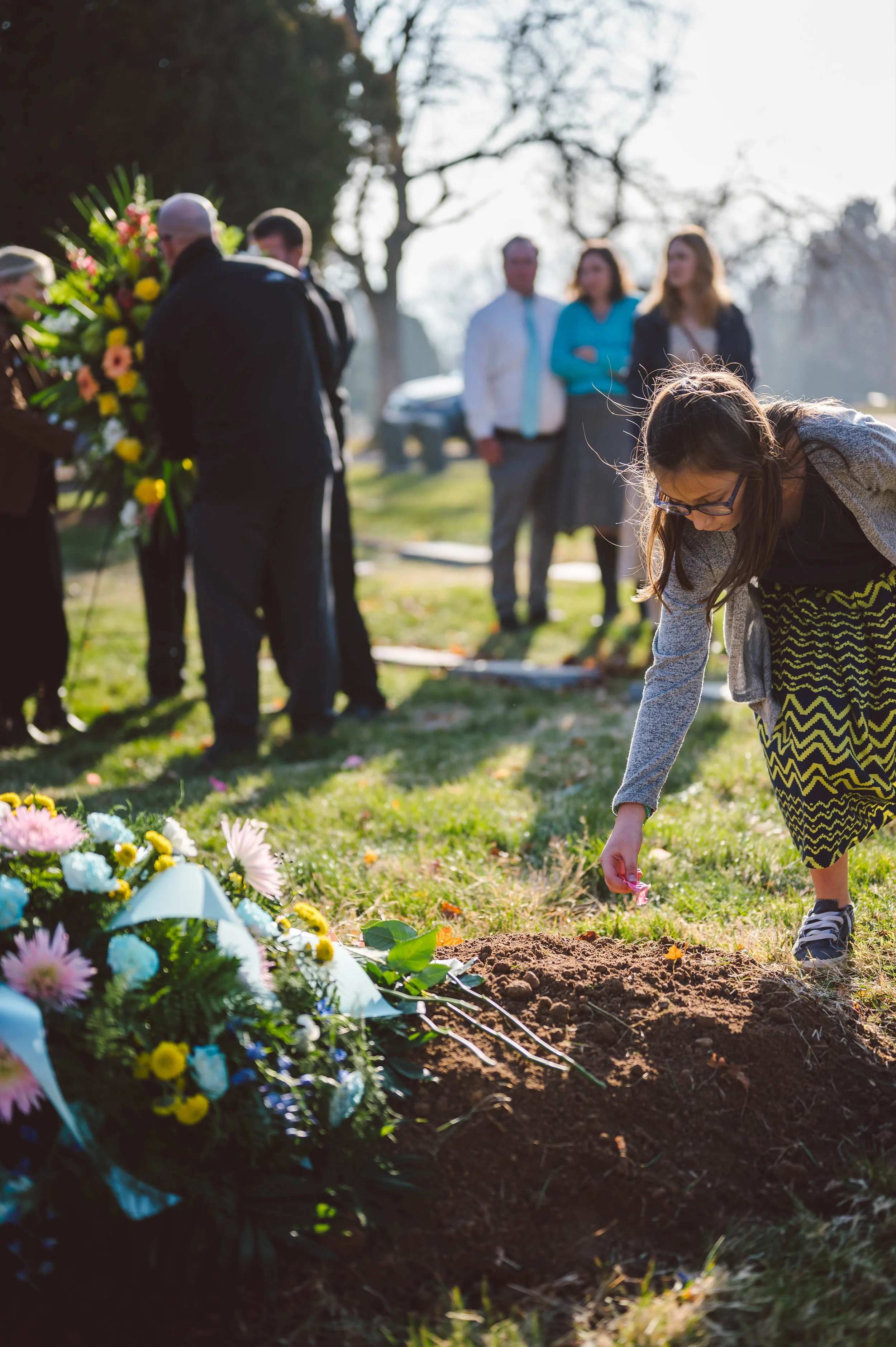 A girl placing a flower on a grave during a funeral or memorial service in a cemetery with other people in the background.