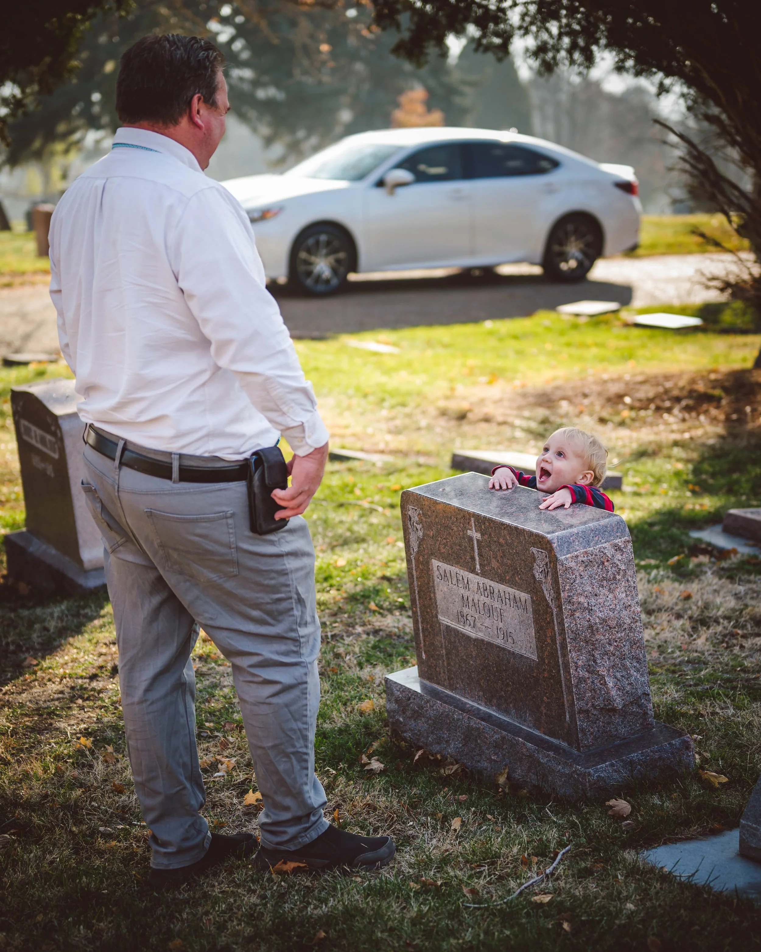 A man in a white shirt and gray pants standing in a cemetery, looking at a young child with blonde hair who is smiling and leaning over a gravestone.