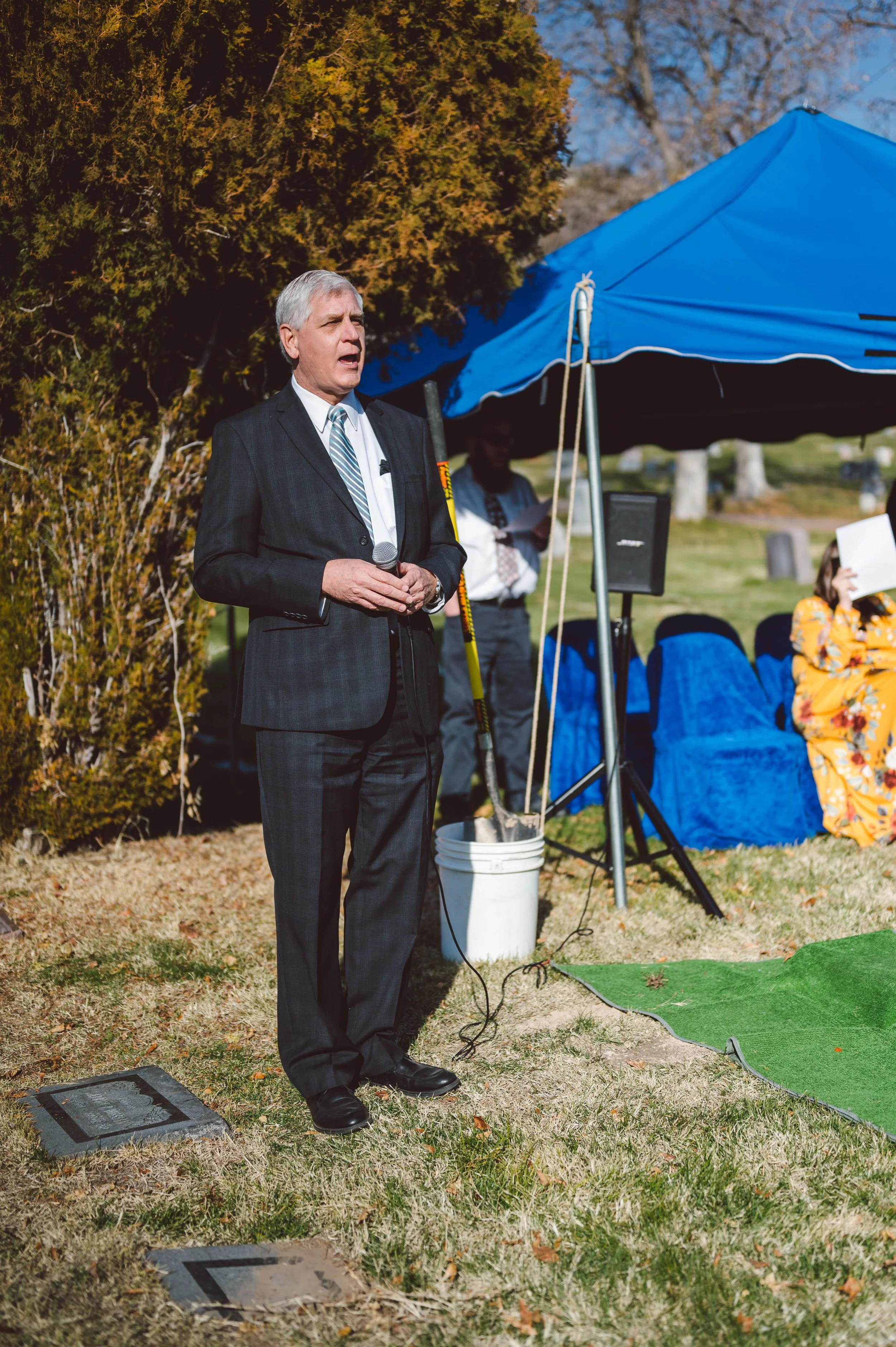 A man in a suit holding a microphone standing outdoors at a memorial site, with a blue canopy and people seated nearby.