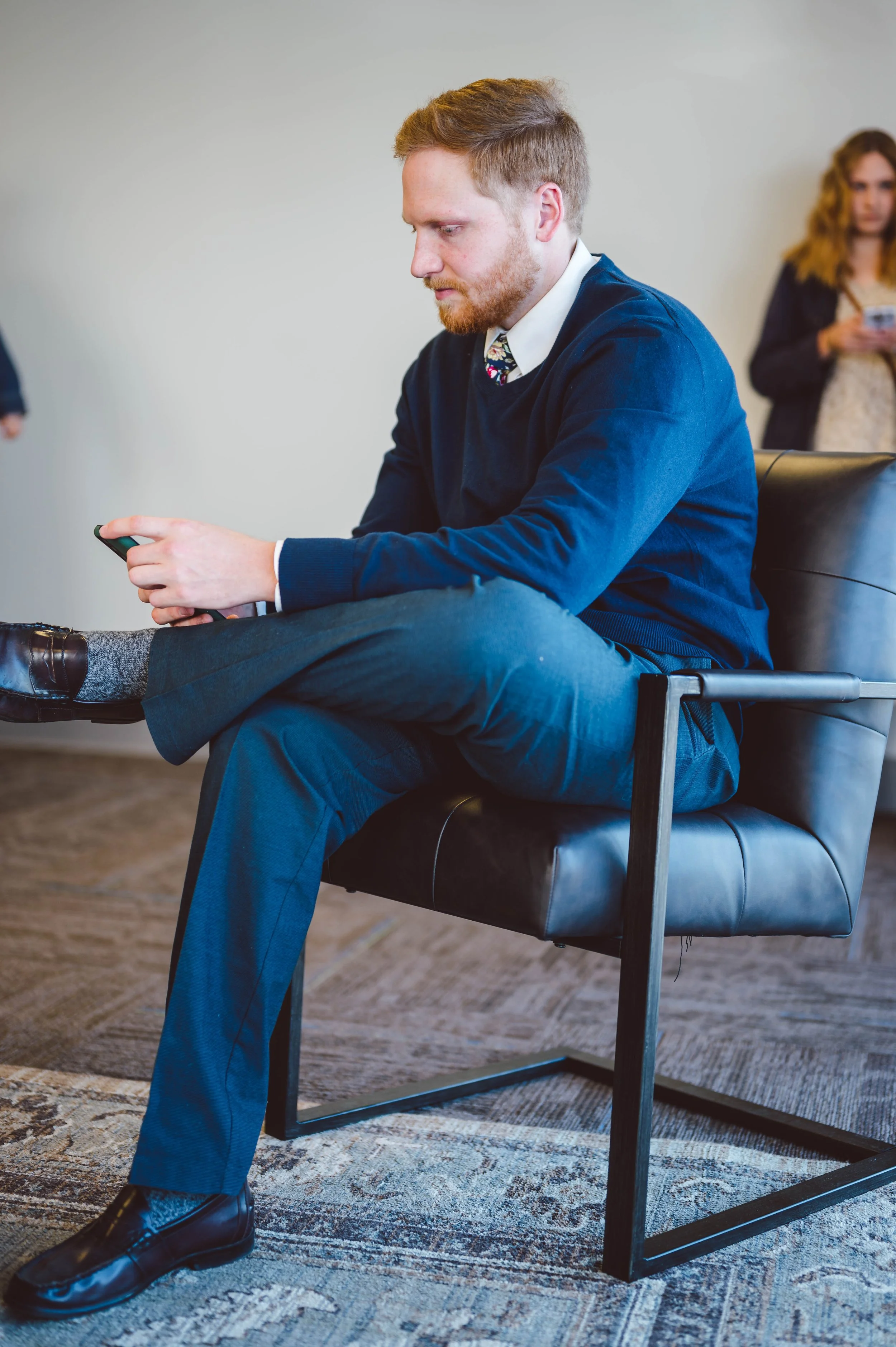 A man with red hair and a beard sitting on a black leather chair, wearing a navy sweater, dress shirt, black pants, and black shoes, looking at his phone. In the background, a woman with long wavy hair is using her phone.