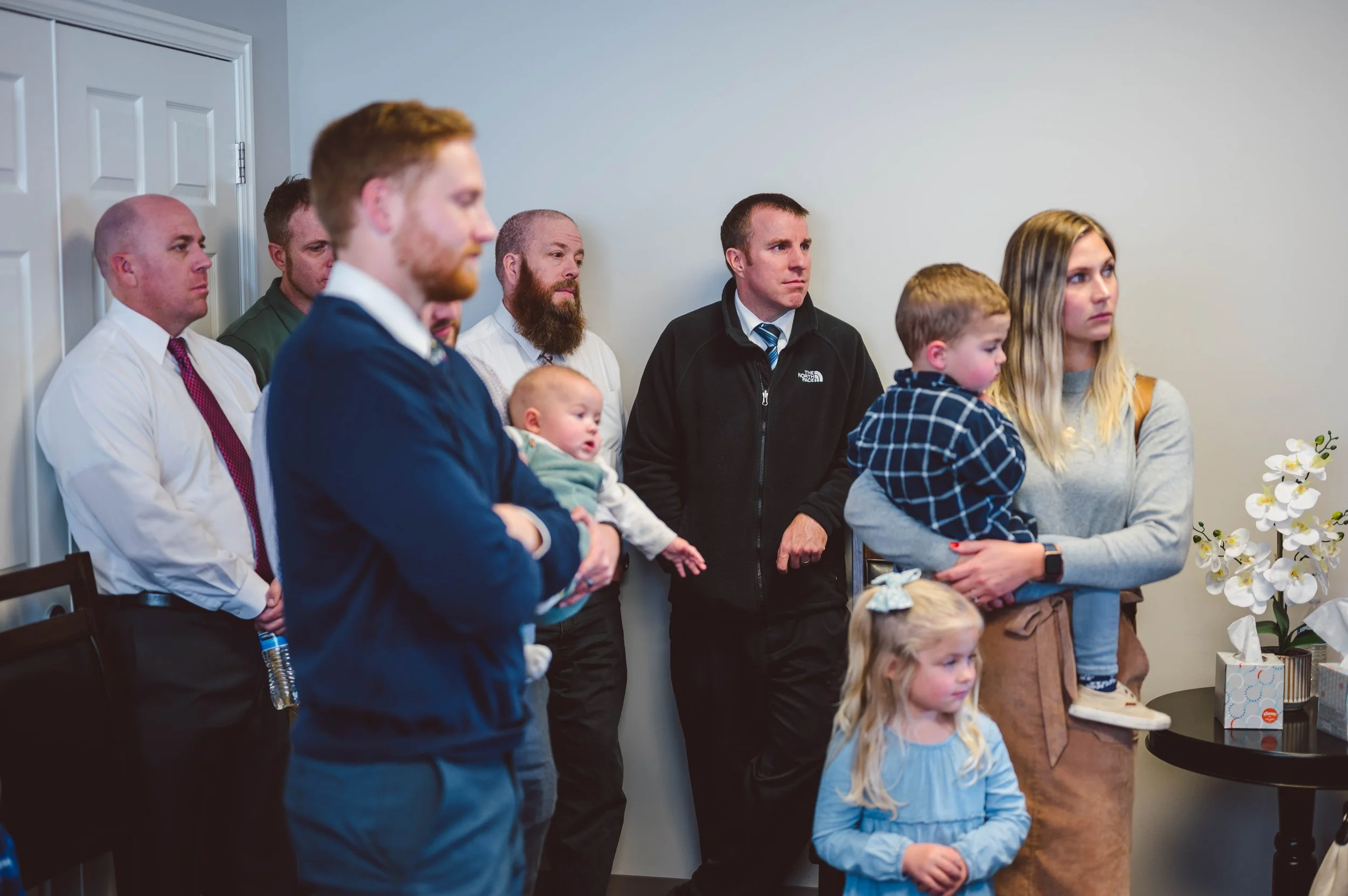 A group of adults and children standing together in a room, likely attending a family or community gathering. They are standing near a wall and a small table with flowers and a tissue box.