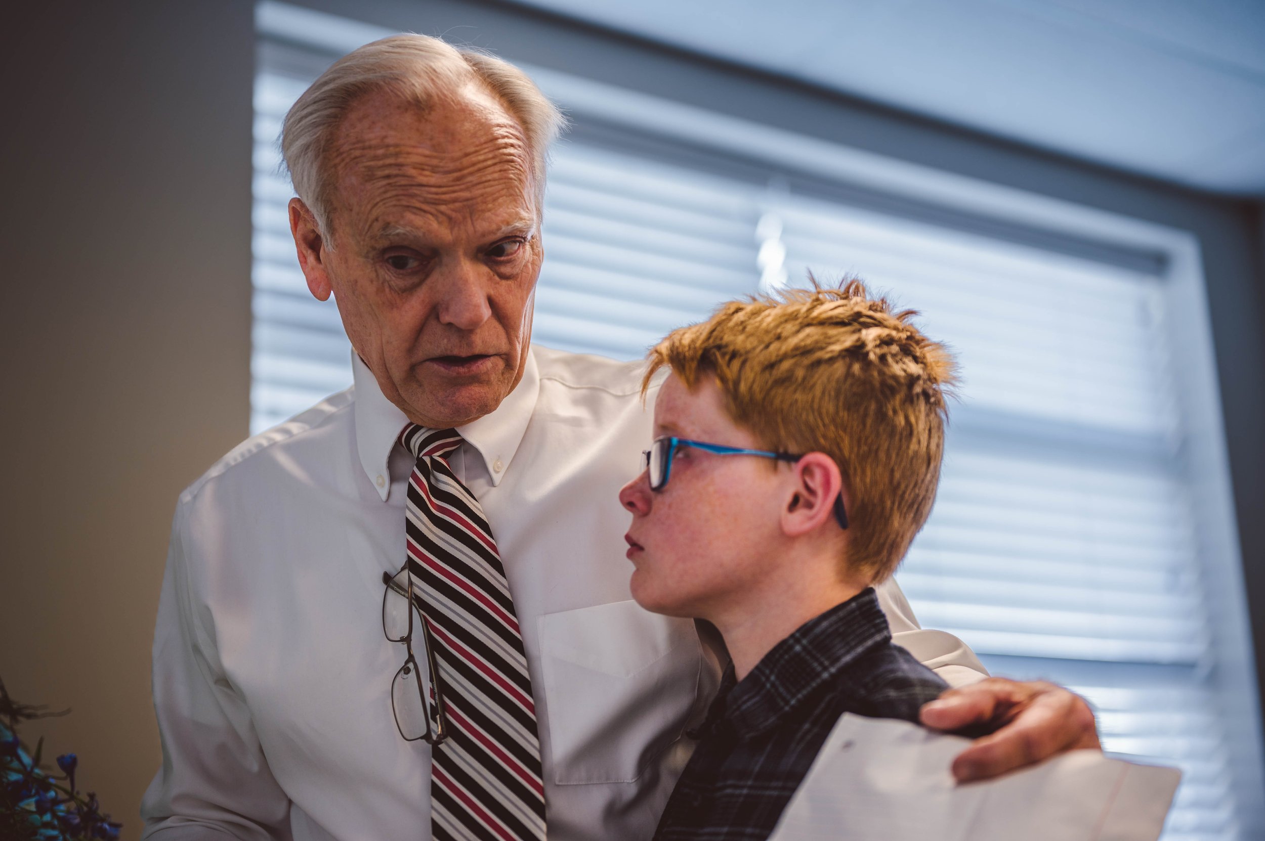An older man dressed in a white shirt and striped tie is comforting a young man with red hair and glasses, wearing a plaid shirt, inside an office with blinds in the background.