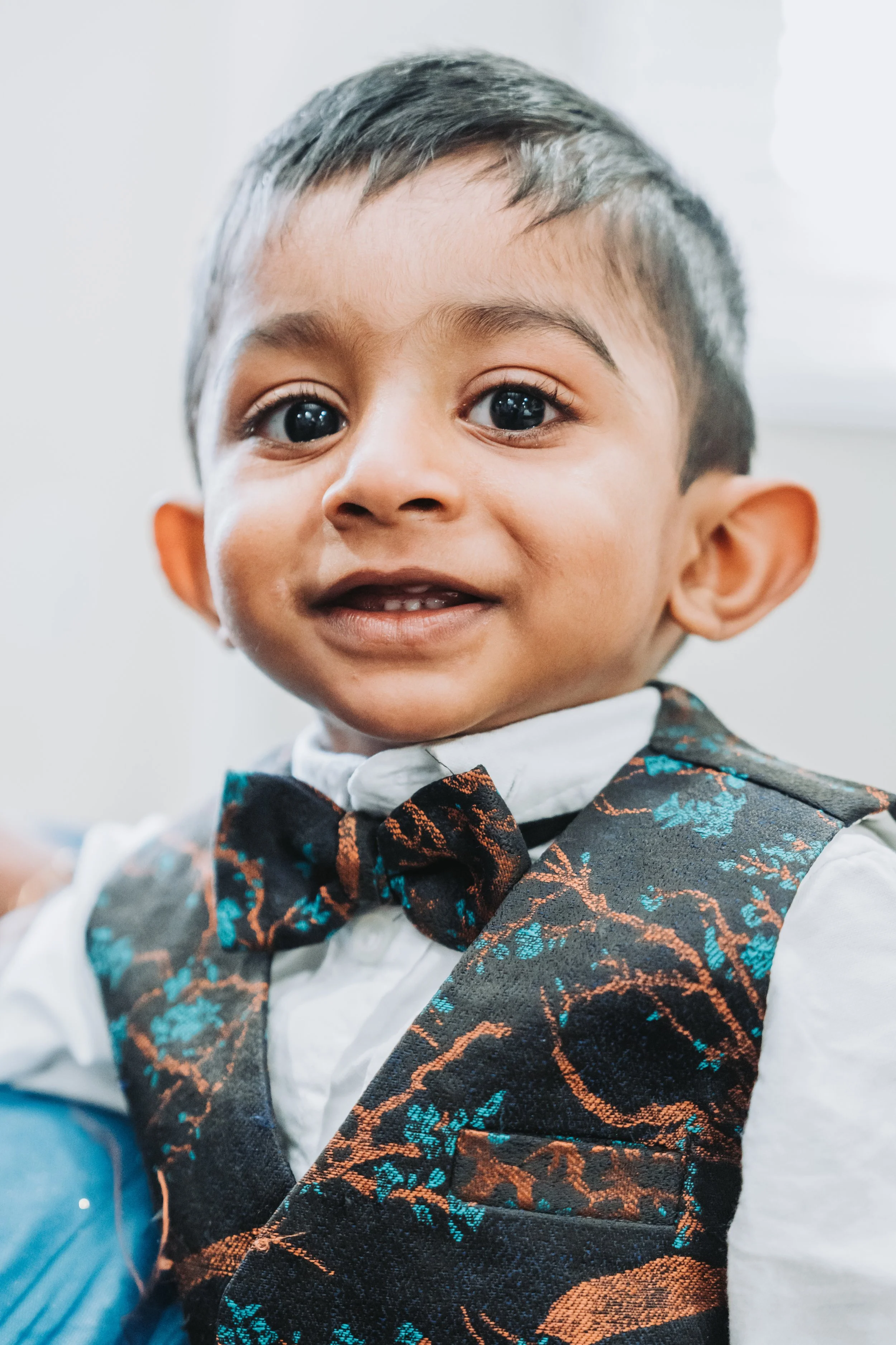 Close-up of a young boy with short dark hair, wearing a white shirt, a black bow tie, and a patterned vest with blue and brown designs.