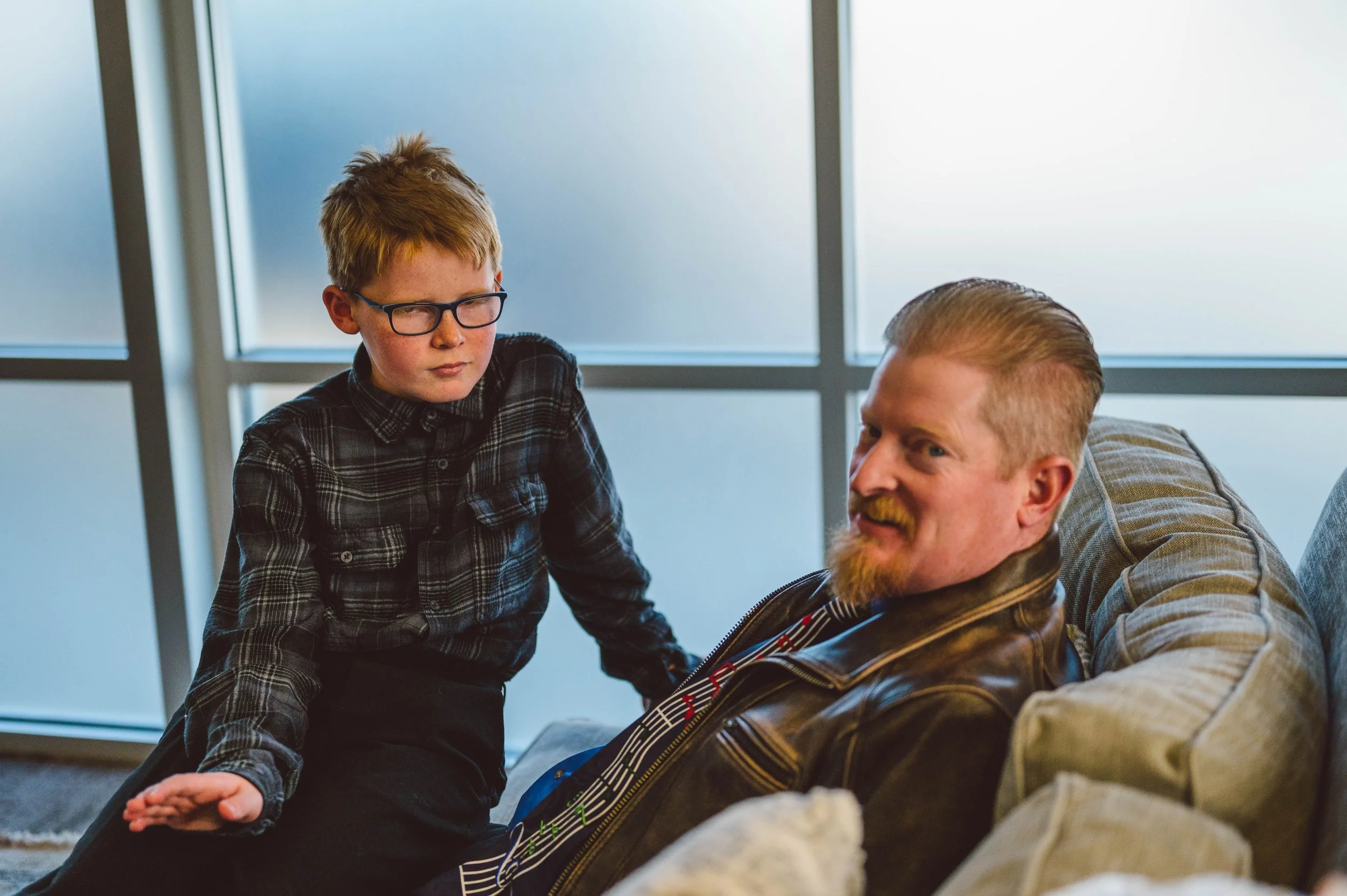 A boy with glasses and a plaid shirt talking to a man with a beard and a leather jacket, sitting on a couch in a modern room with windows.