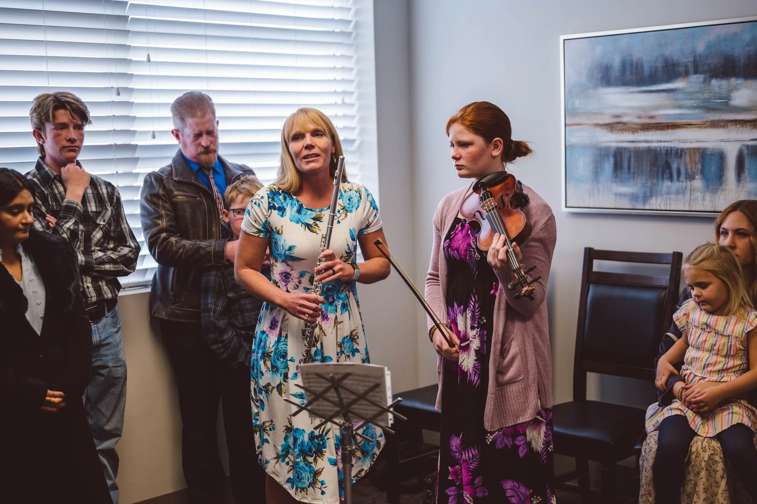 A group of people gathered indoors, with two women holding musical instruments, a flute and a violin, standing in front of a window with blinds. Others are watching, including a young girl sitting on a chair to the right.