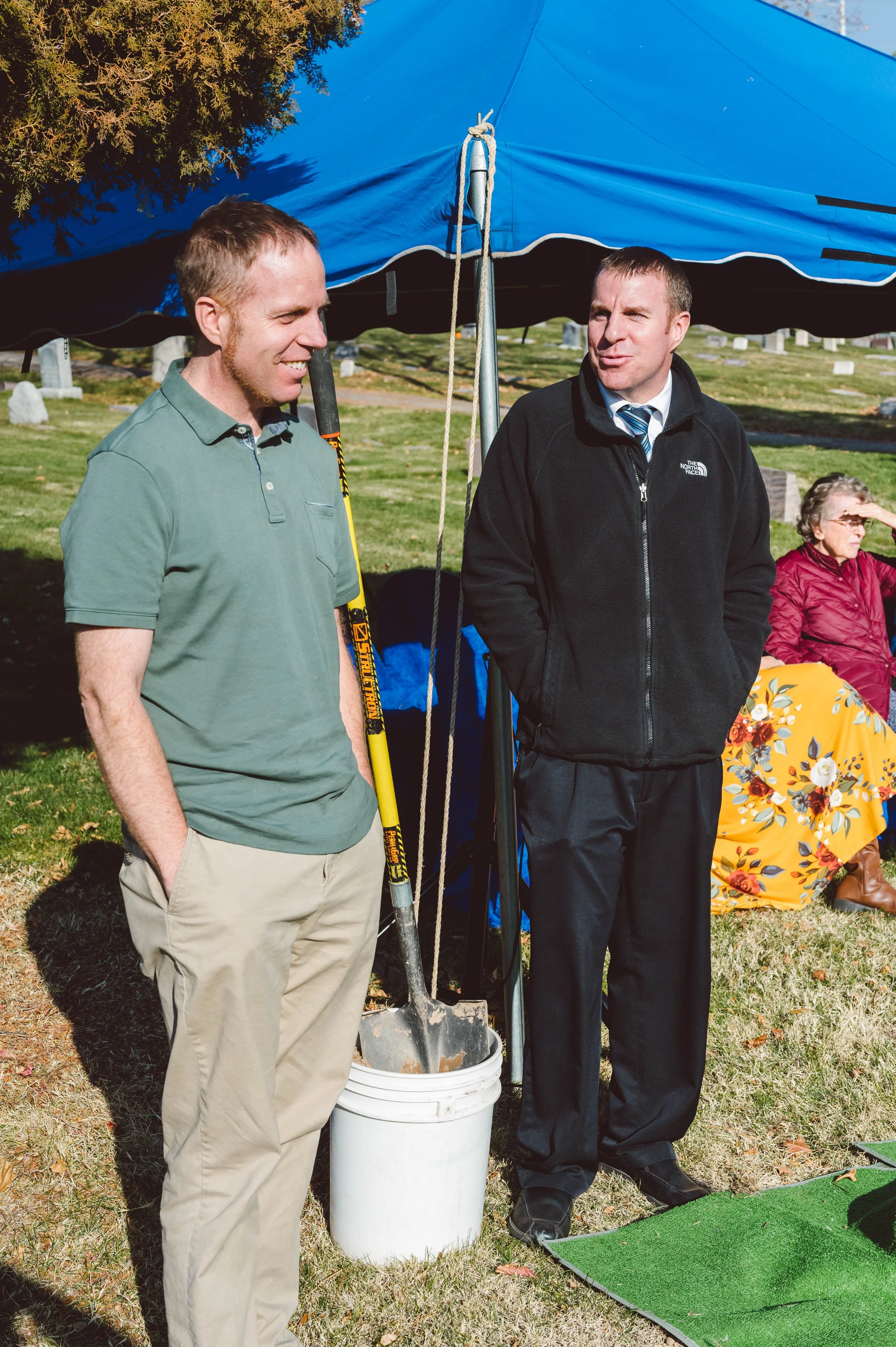 Two men are standing outdoors under a blue tent, engaged in conversation. One man is holding a shovel and smiling, while the other is dressed in a black jacket and surrounded by grave markers, indicating a cemetery setting. A woman sitting nearby is 