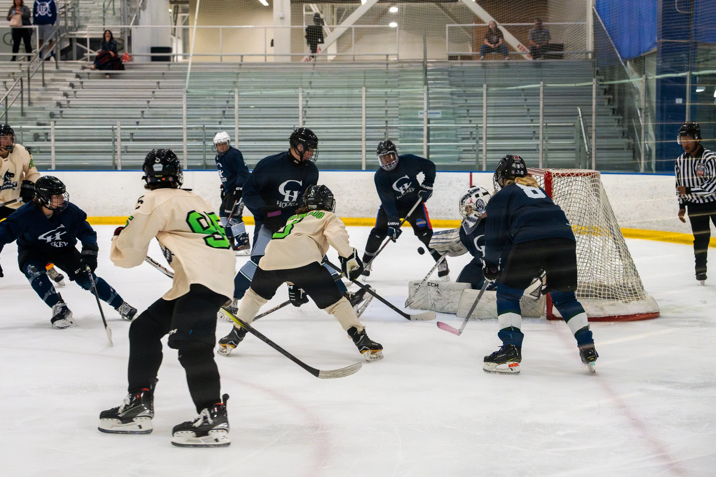A group of ice hockey players, wearing jerseys and helmets, are engaged in a game around the goal. The goaltender is crouched in front of the net, attempting to block the puck, while other players are trying to score or defend.