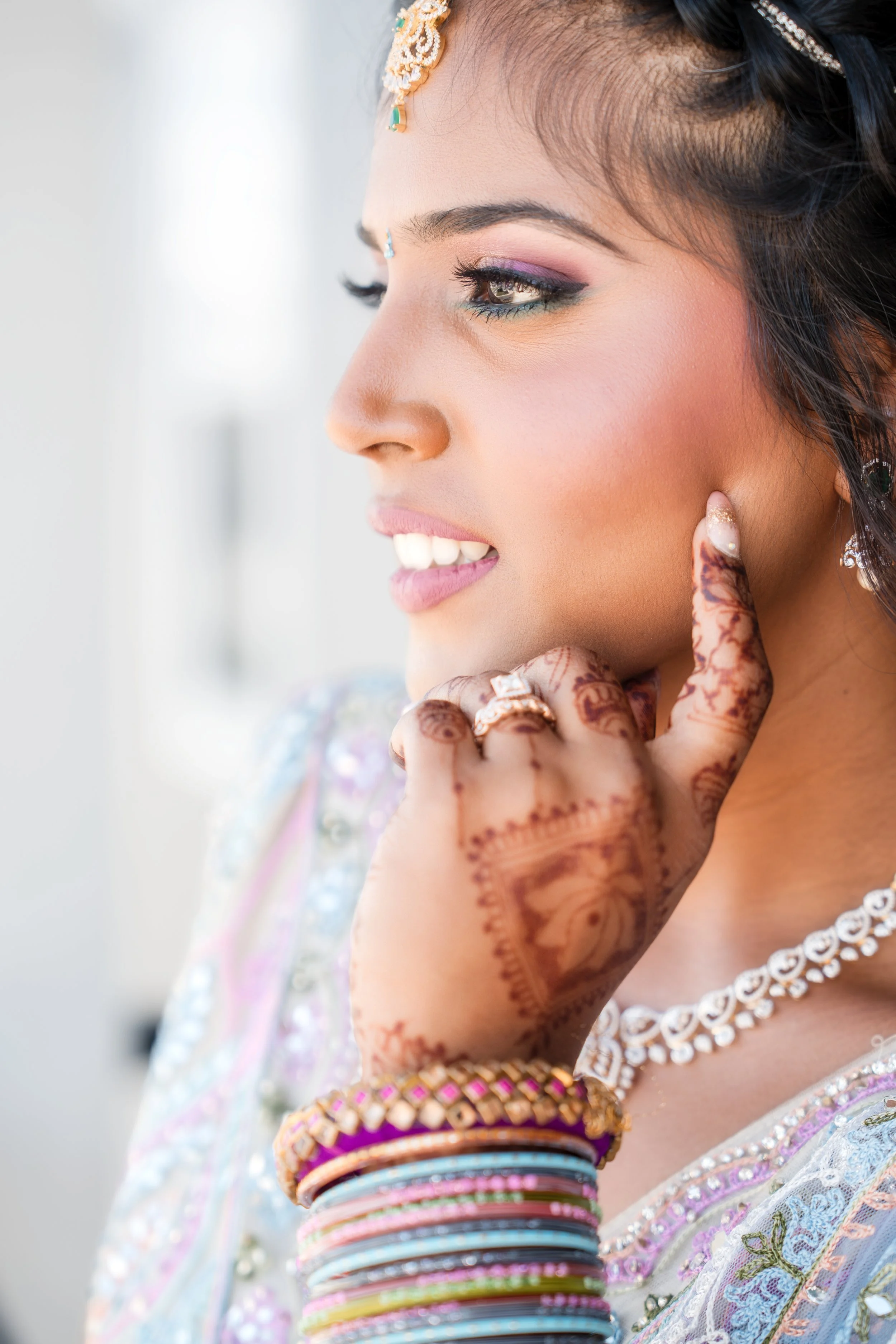 A young woman dressed in traditional South Asian attire, wearing jewelry, mehndi on her hand, and colorful bangles, looking to the side with her hand near her face.