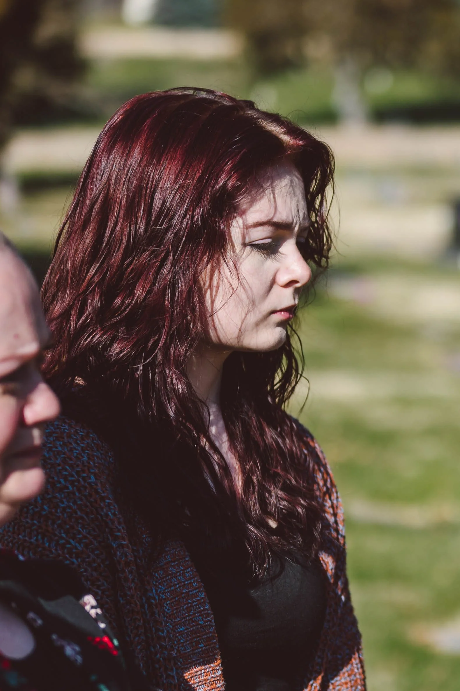 A woman with red wavy hair is standing outdoors with a somber expression, wearing a dark top and a patterned sweater.