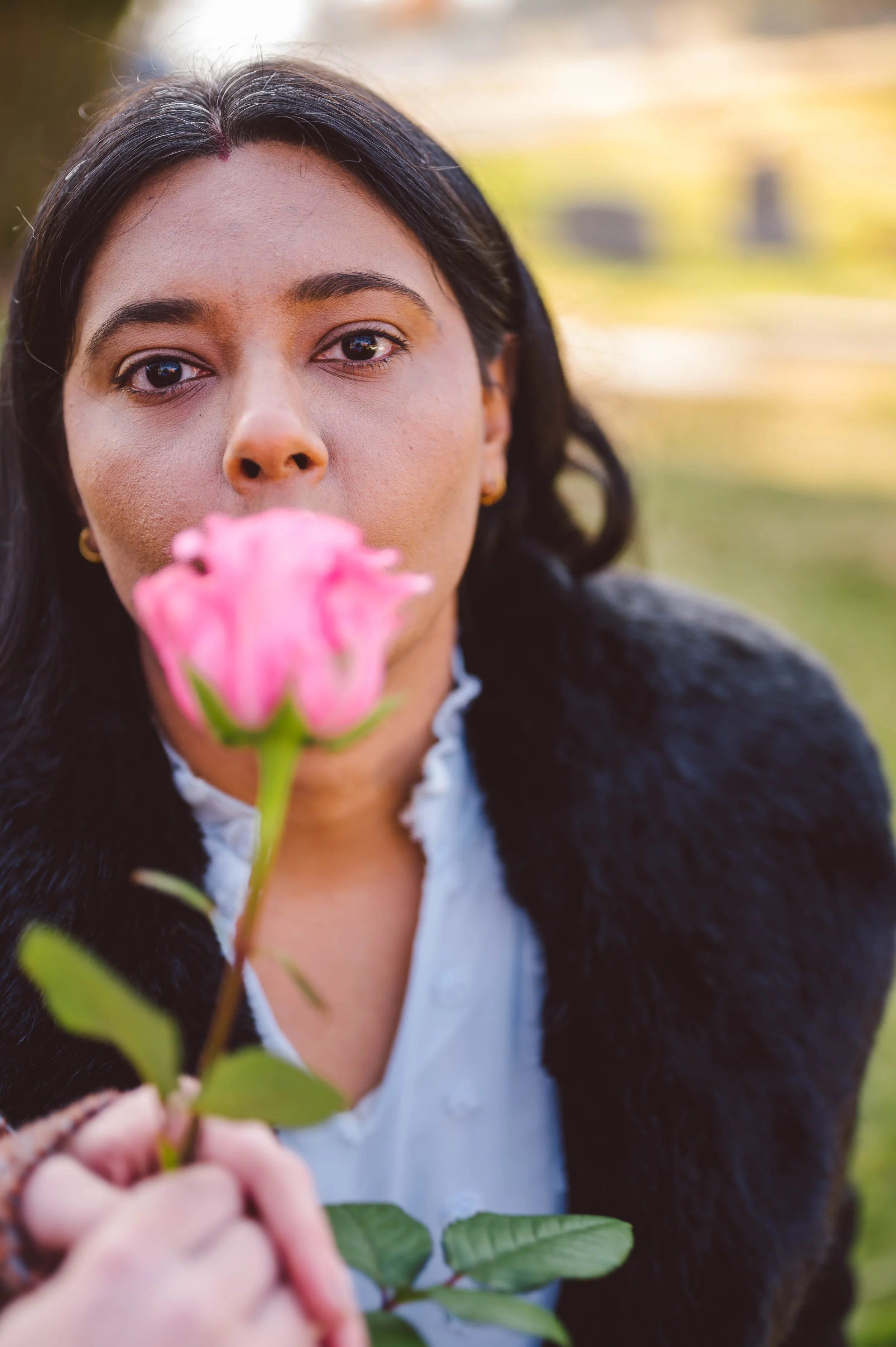 A woman with dark hair holding a pink rose close to her face outdoors, with a blurred background of grass and trees.