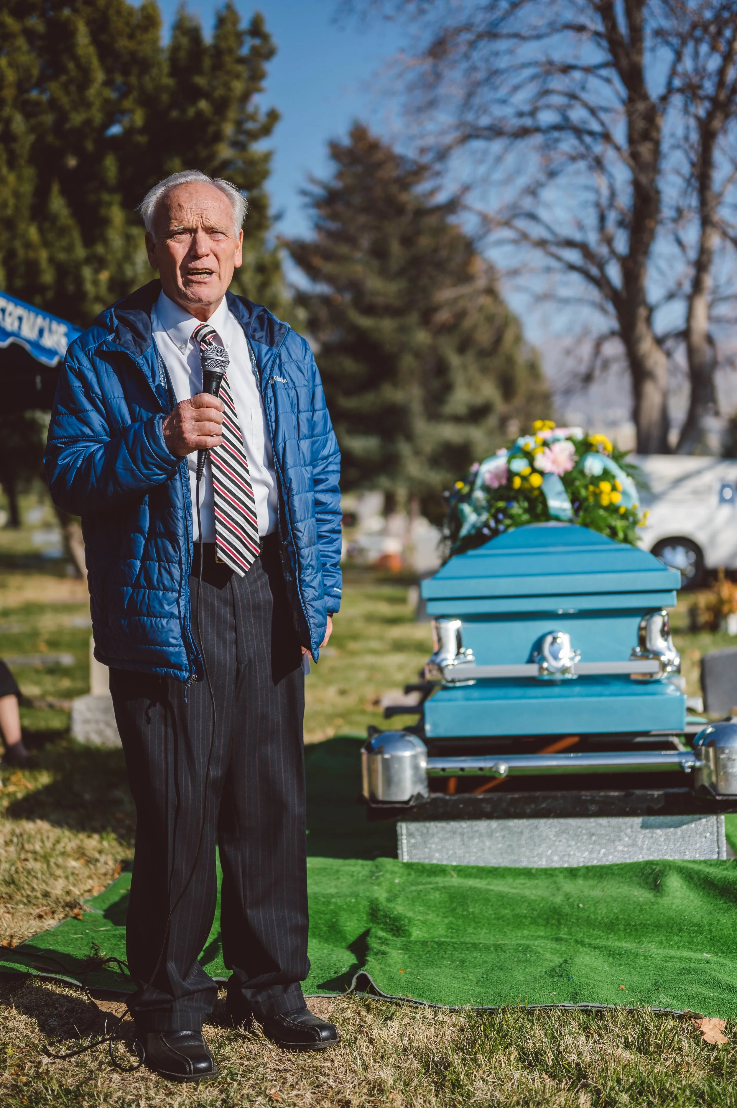 An elderly man in a blue jacket, white shirt, and striped tie speaking into a microphone at a graveside funeral service during daytime. A blue coffin with a floral arrangement on top is nearby, and the setting is an outdoor cemetery with trees and ca