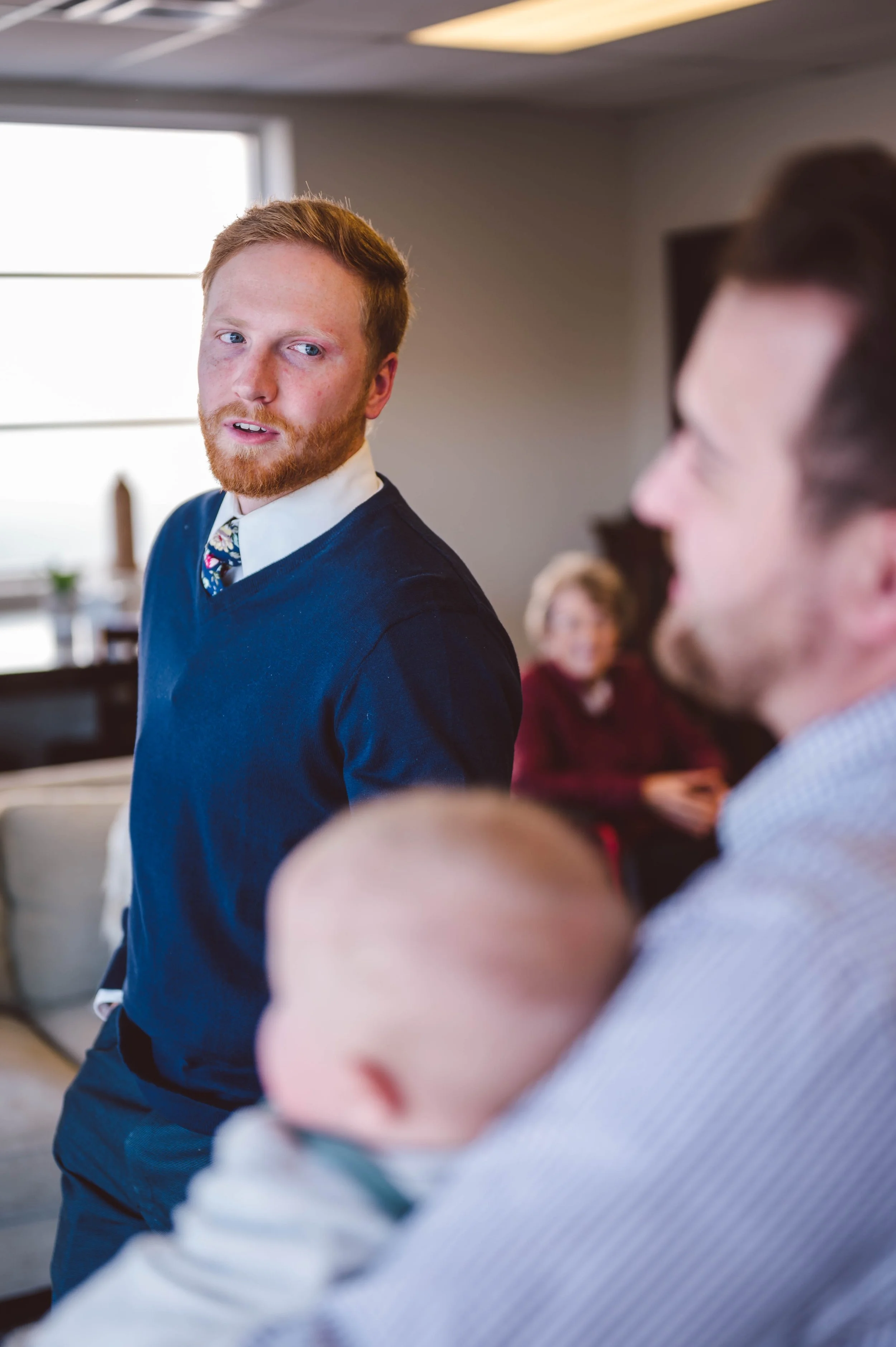 A young man with red hair and a beard, dressed in a white shirt, navy sweater, and patterned tie, is speaking to a person holding a baby. An older woman with glasses and short blonde hair is blurred in the background in a room with natural light comi
