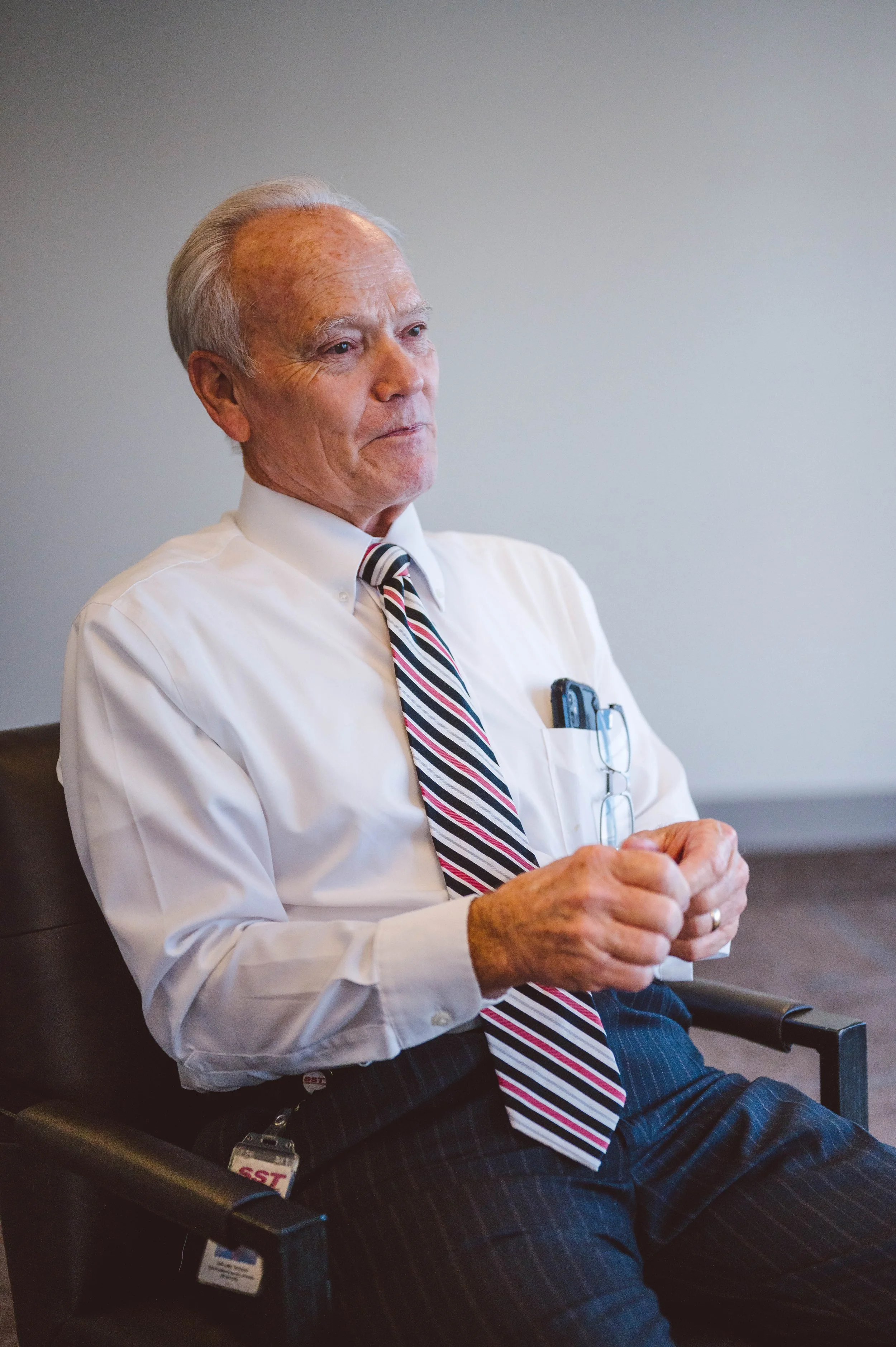 An elderly man with gray hair wearing a white dress shirt, striped tie, and dark pants sitting in a black chair with his hands clasped together, in a professional setting.