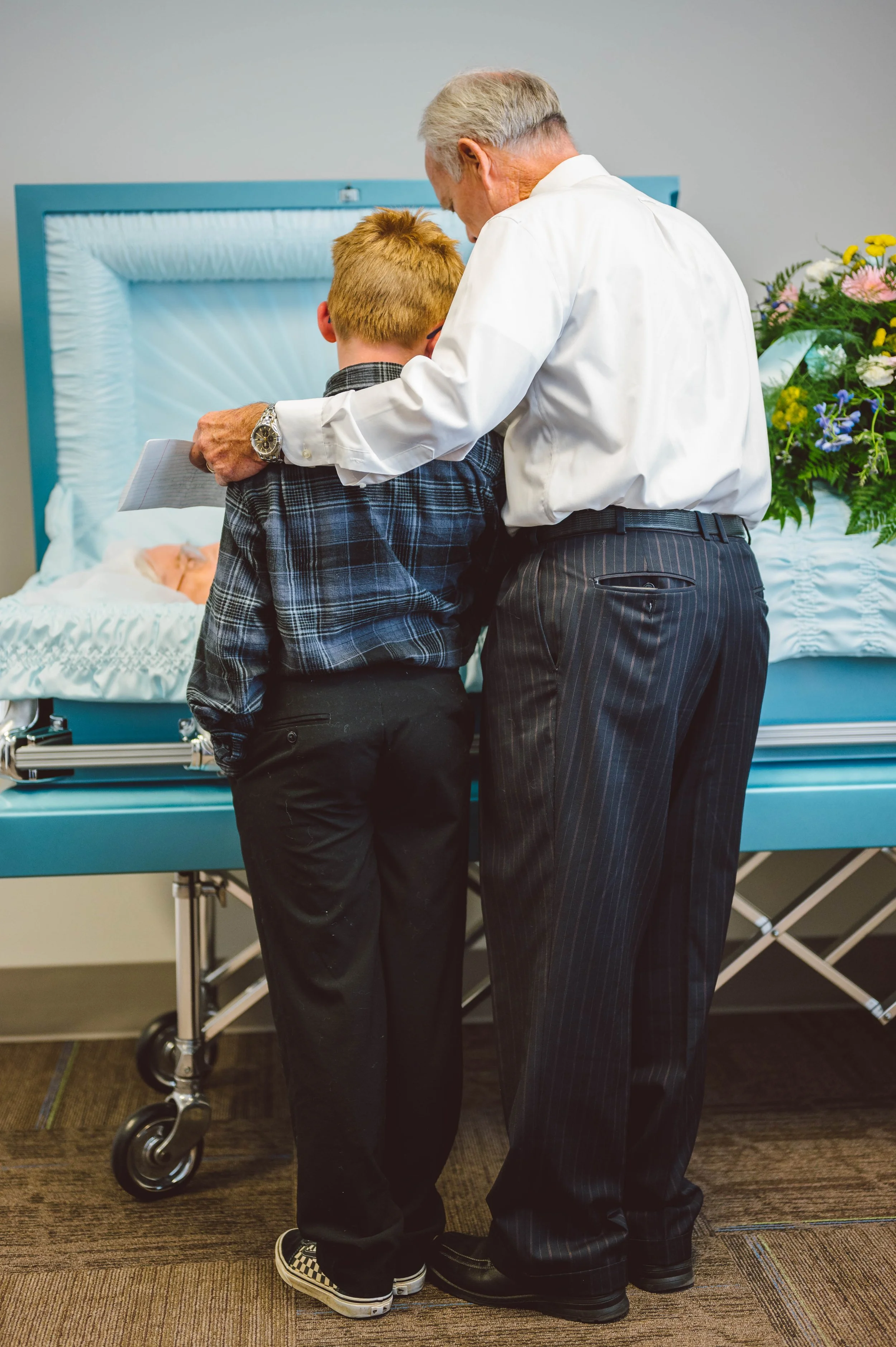 An older man and a young boy mourn at a cremation service, standing close together with the casket in front of them and feeling emotional.