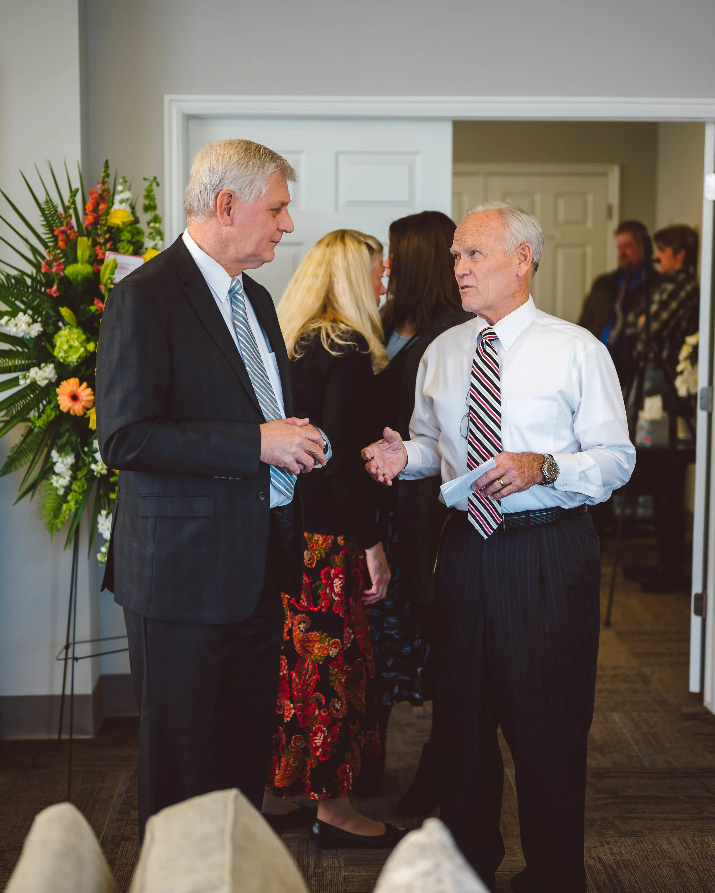 Two elderly men wearing suits and ties having a conversation at a social event, with other people in the background and a large floral arrangement nearby.