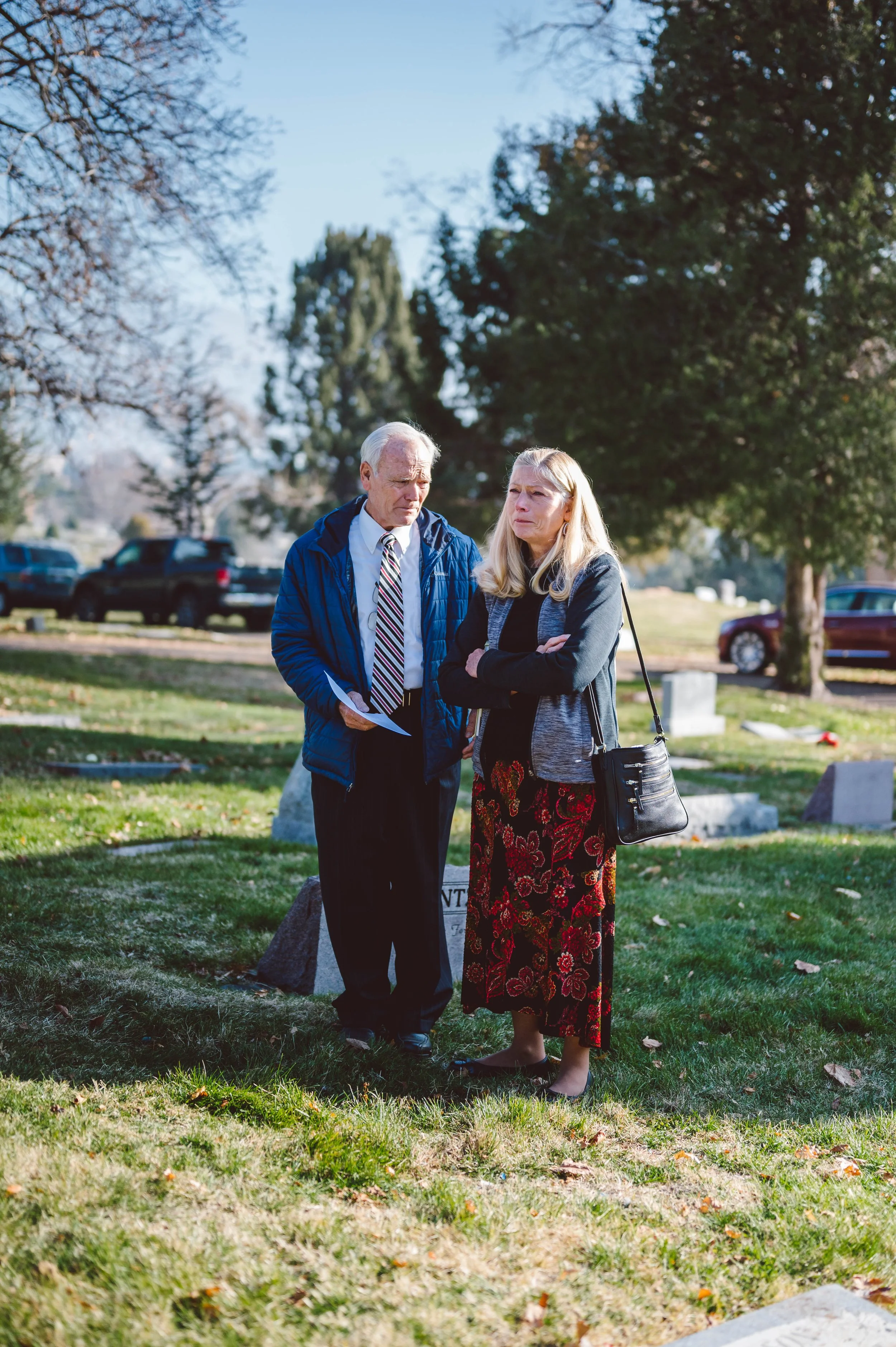 A man and woman mourning at a cemetery, standing by a gravestone on a grassy area with trees and parked cars in the background.