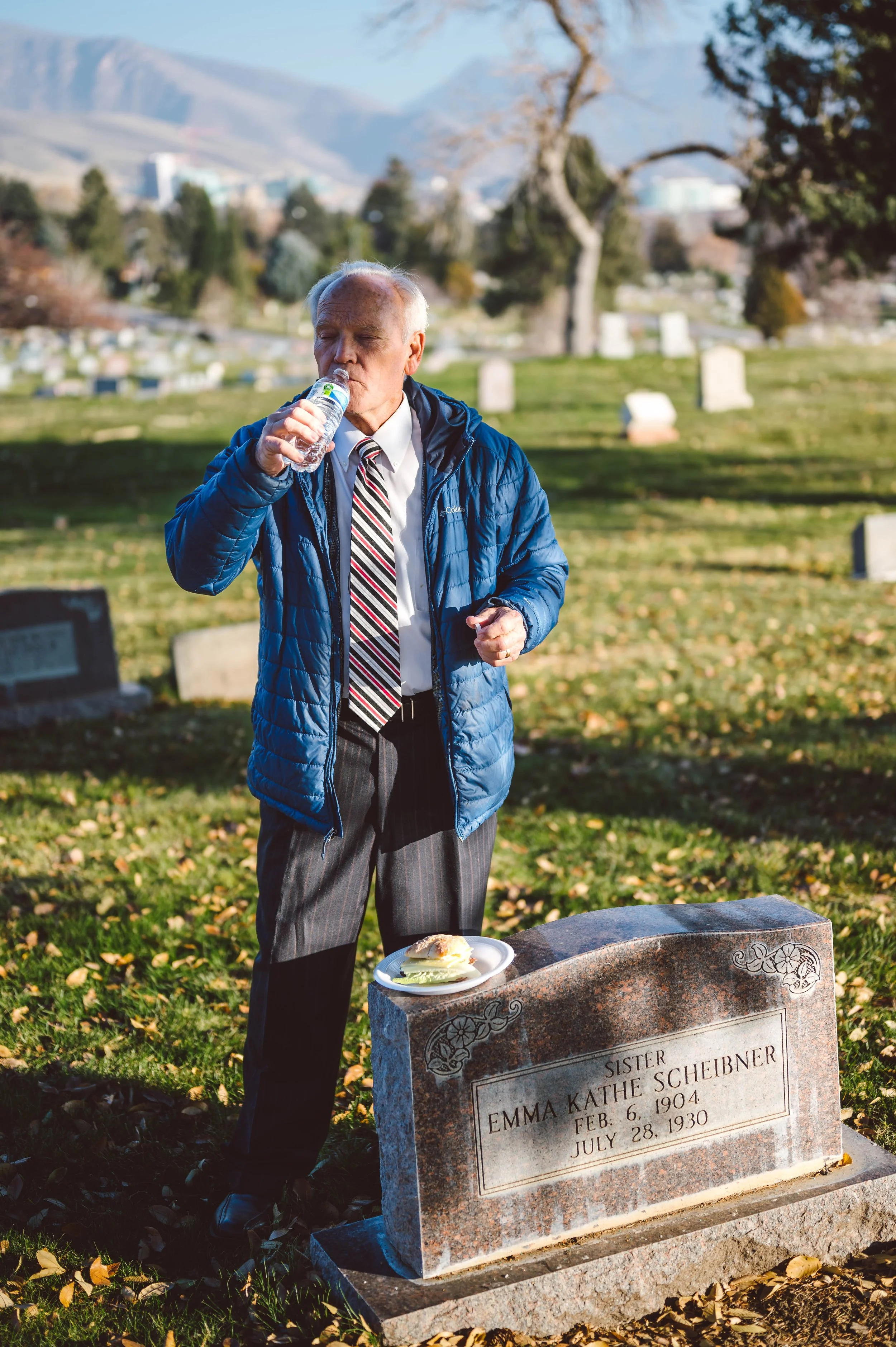 A man in a blue jacket, dress shirt, and striped tie stands next to a gravestone, drinking water from a bottle, with a plate of food on the gravestone, in a cemetery with trees and mountains in the background.