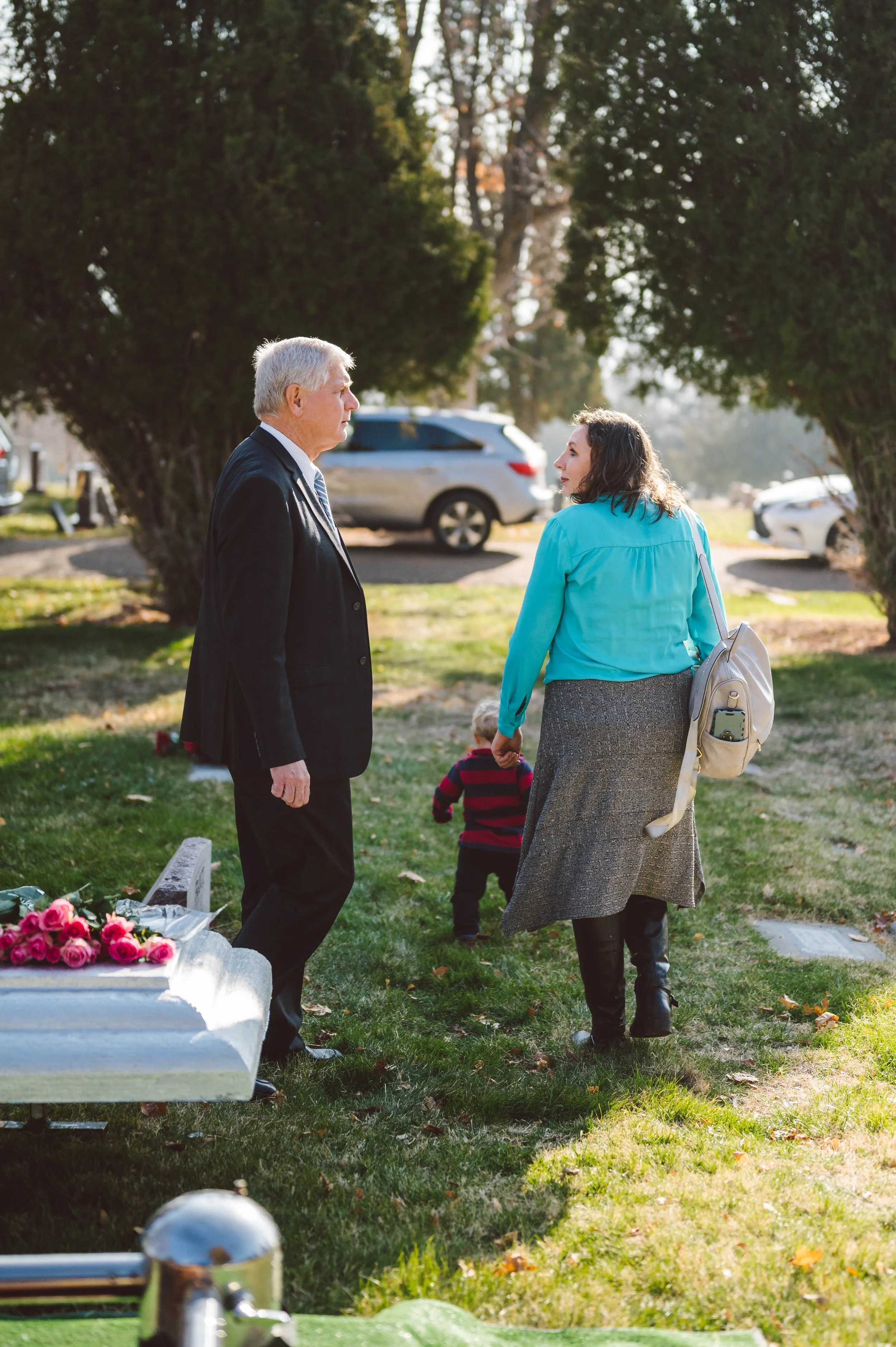 A man and woman holding hands and talking in a park-like setting with trees and parked cars in the background; a small child is walking away from the camera.