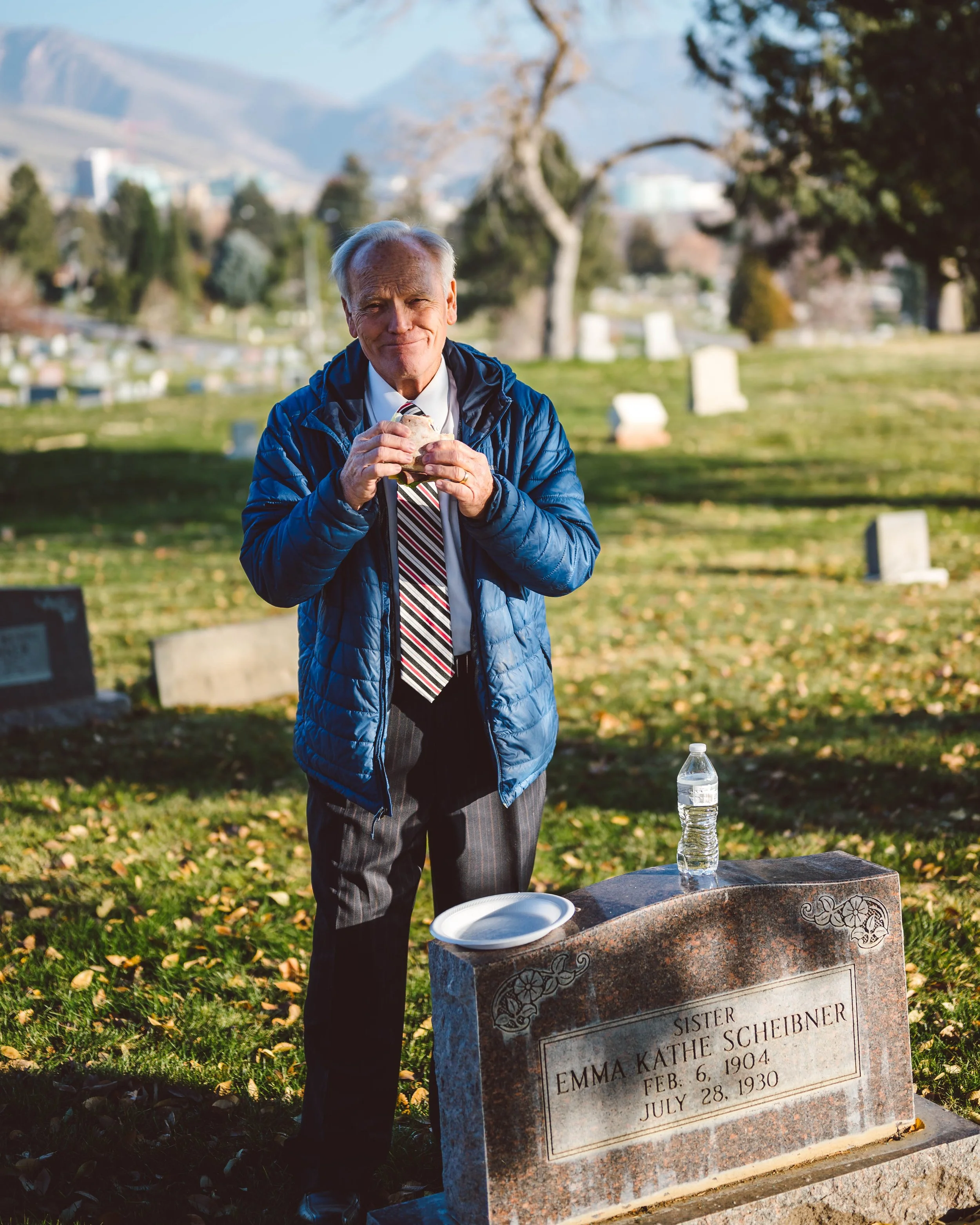 An elderly man in a blue puffy jacket and a striped tie standing at a graveyard holding a cookie. A water bottle and a paper plate are on the tombstone, which reads 'Emma Kathe Scheibner, Feb. 6, 1904 - July 28, 1930.'