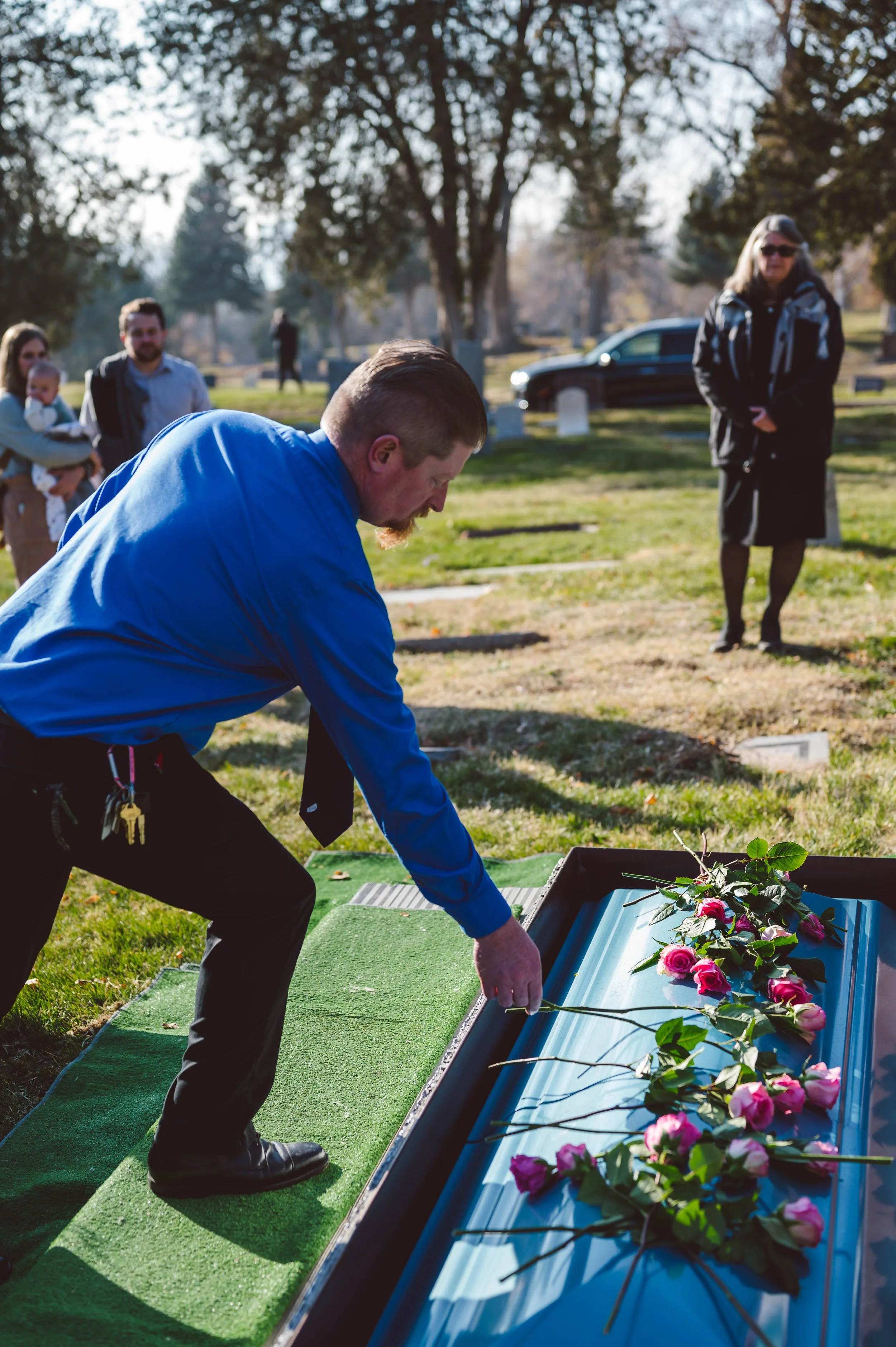 Man in blue shirt laying flowers on a grave at a cemetery during daytime, surrounded by other mourners.