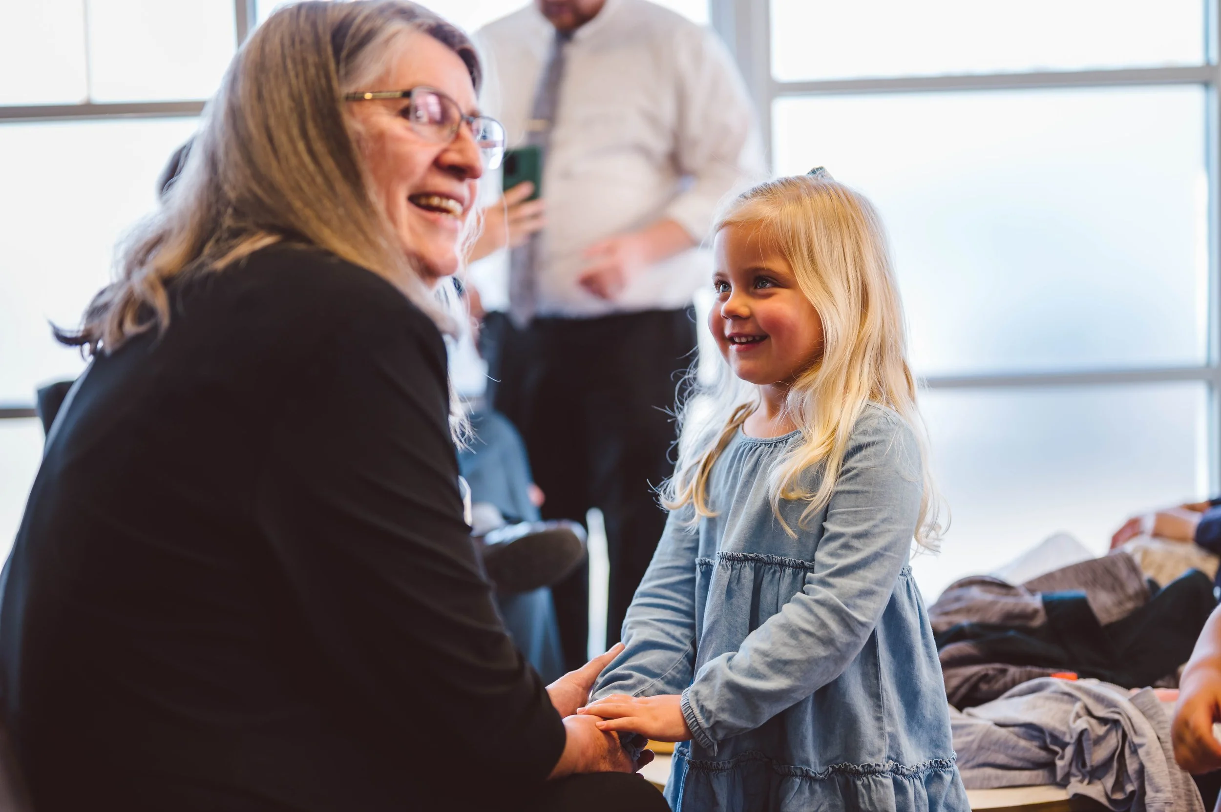 A woman with glasses and a black shirt is smiling and talking to a young girl with long blonde hair, who is wearing a long-sleeved blue dress. They are indoors, near large windows, with people and clothes visible in the background.