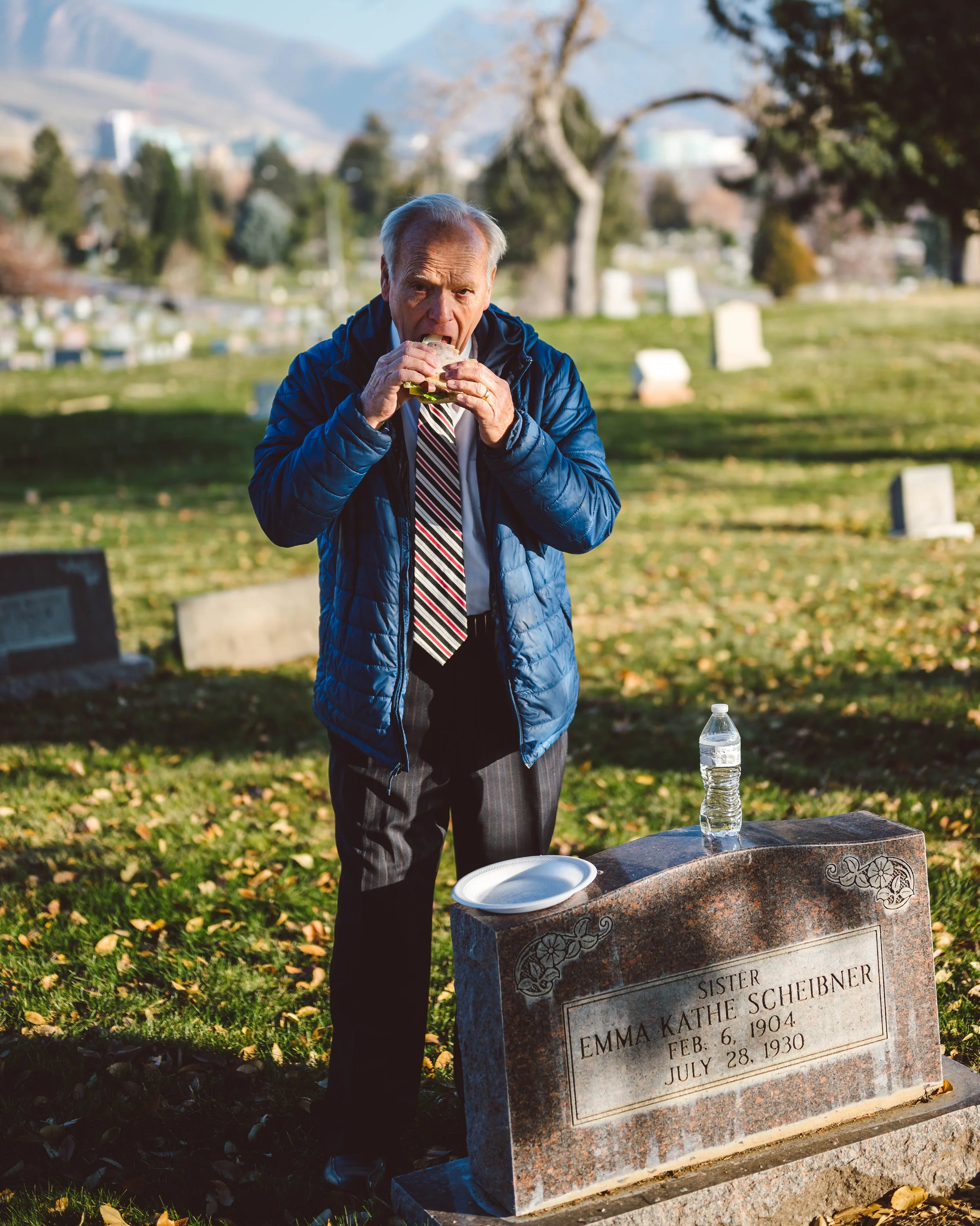 An elderly man in a blue jacket, striped tie, and dark pants standing in a cemetery, biting into a sandwich near a tombstone with a water bottle and paper plate on it, surrounded by gravestones and trees.