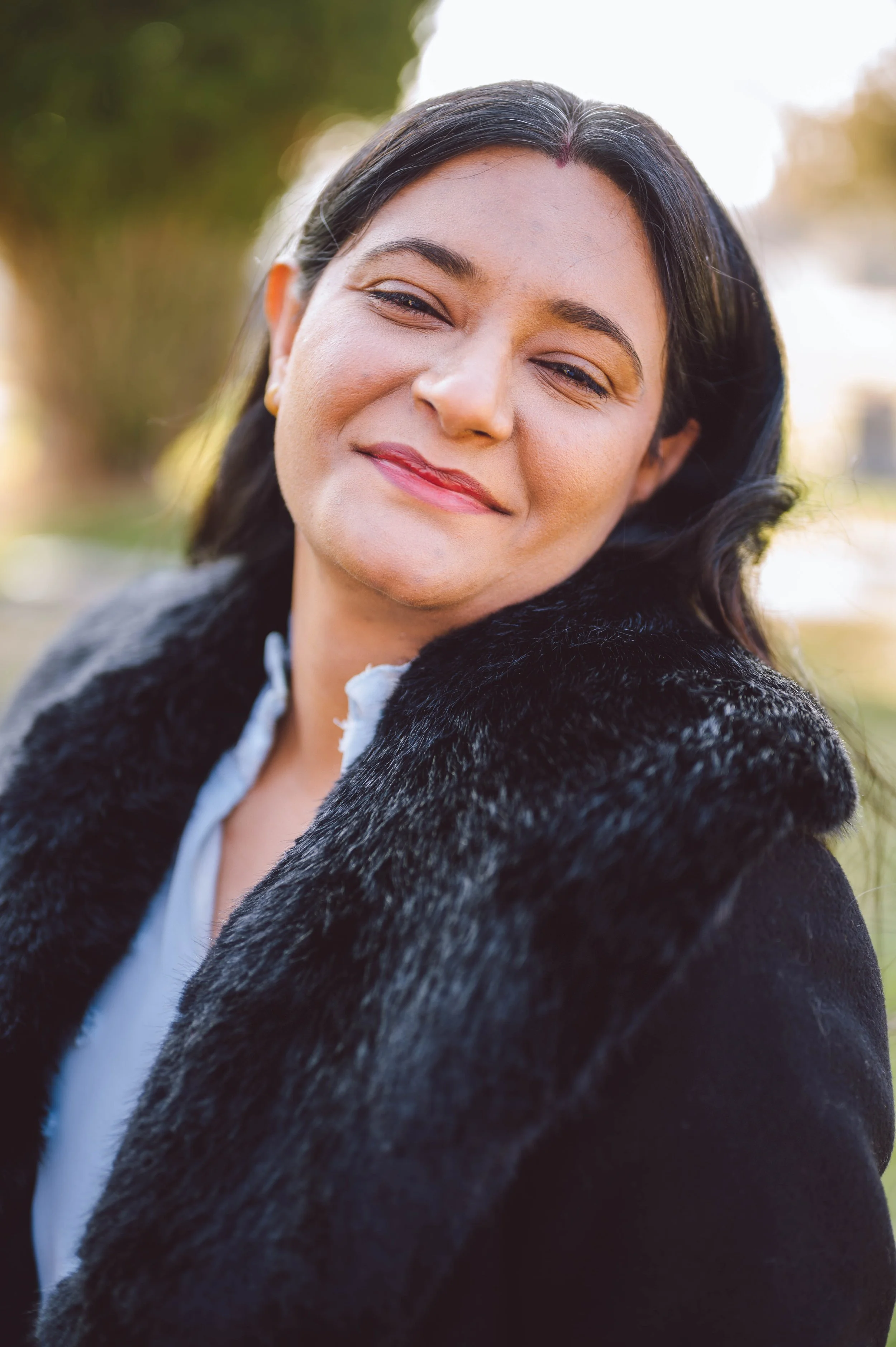 A woman with dark hair smiling and winking outdoors, wearing a black fur coat and white shirt.