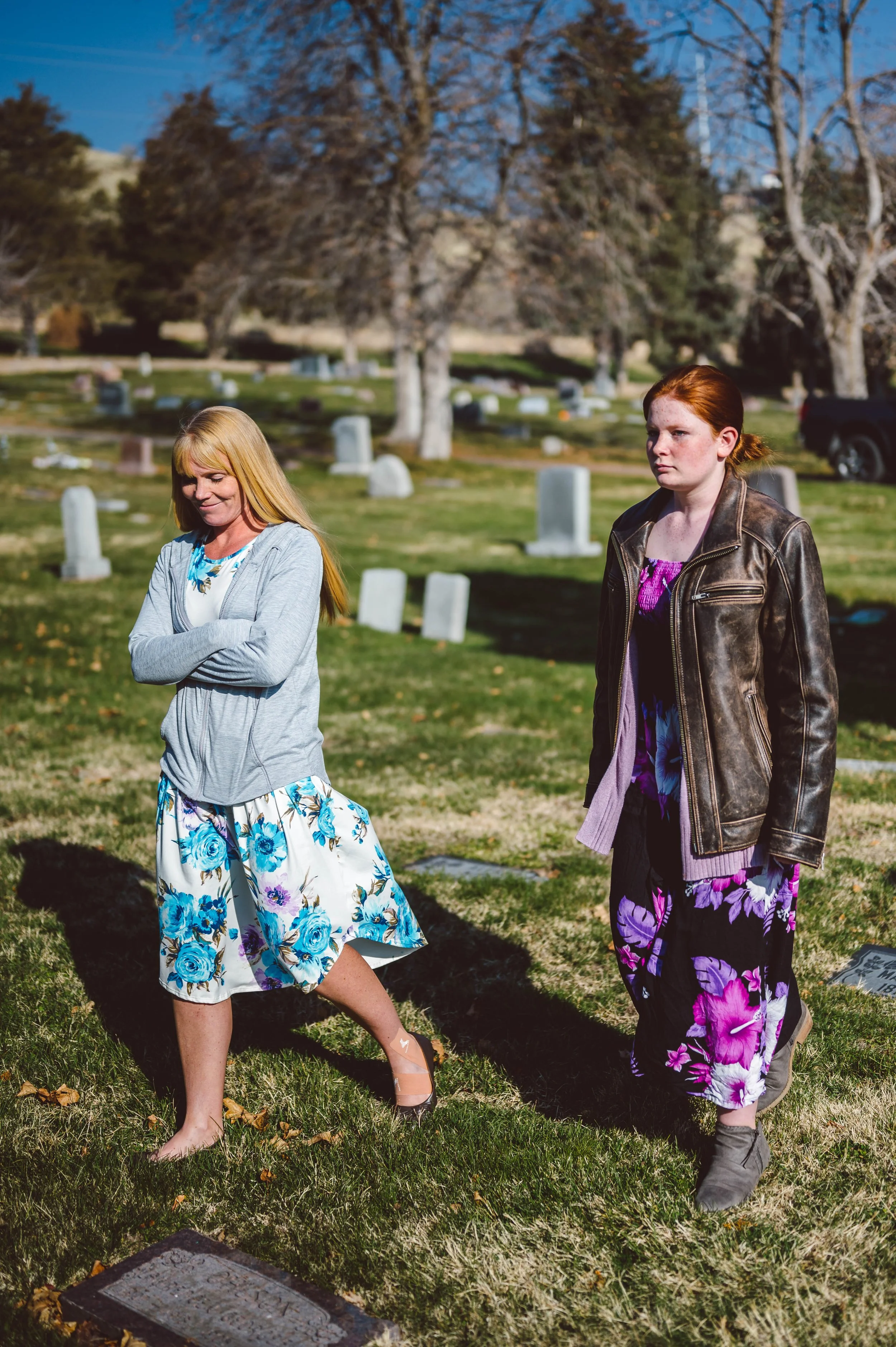 Two women standing in a cemetery with headstones and leafless trees in the background. One woman has blonde hair, is dressed in a floral skirt and gray cardigan, and is smiling with her eyes closed. The other woman has red hair, is wearing a leather 