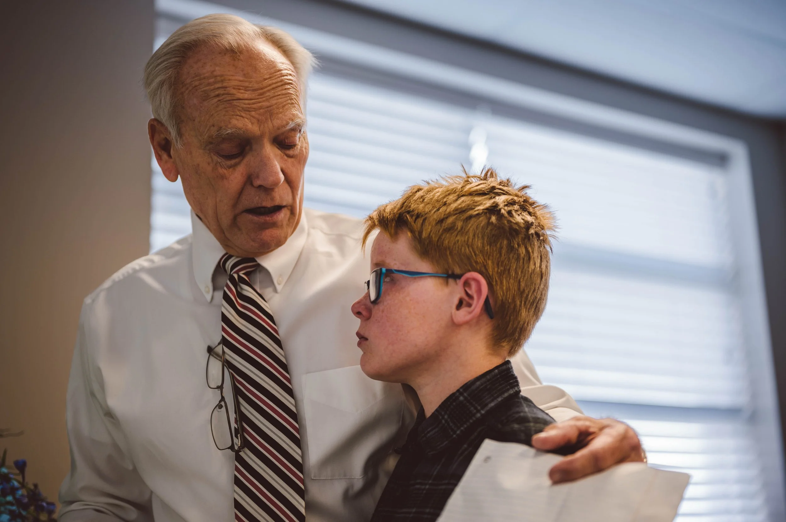 An elderly man comforting a young man with red hair and glasses in a medical setting.