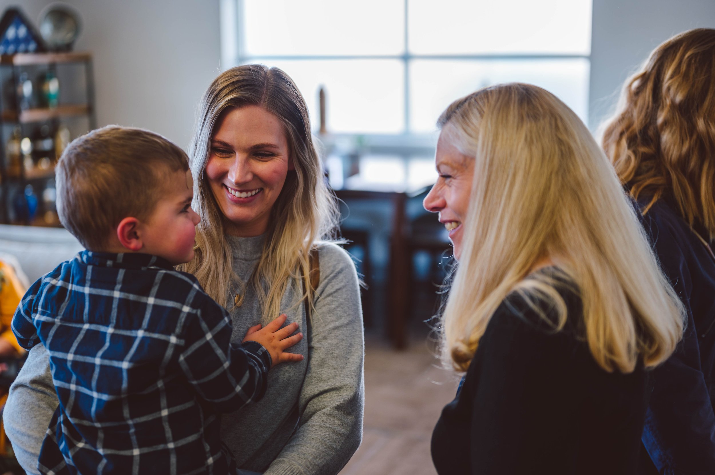 Three women and a young boy smiling and talking in a cozy indoor setting with large windows and shelves in the background.
