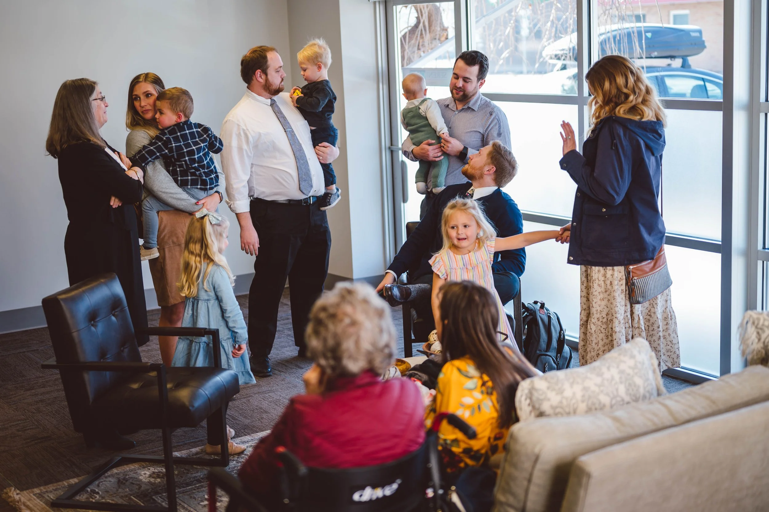 A group of adults and children gathered in a bright room near large windows, with some seated and some standing, engaging in conversation and interacting.