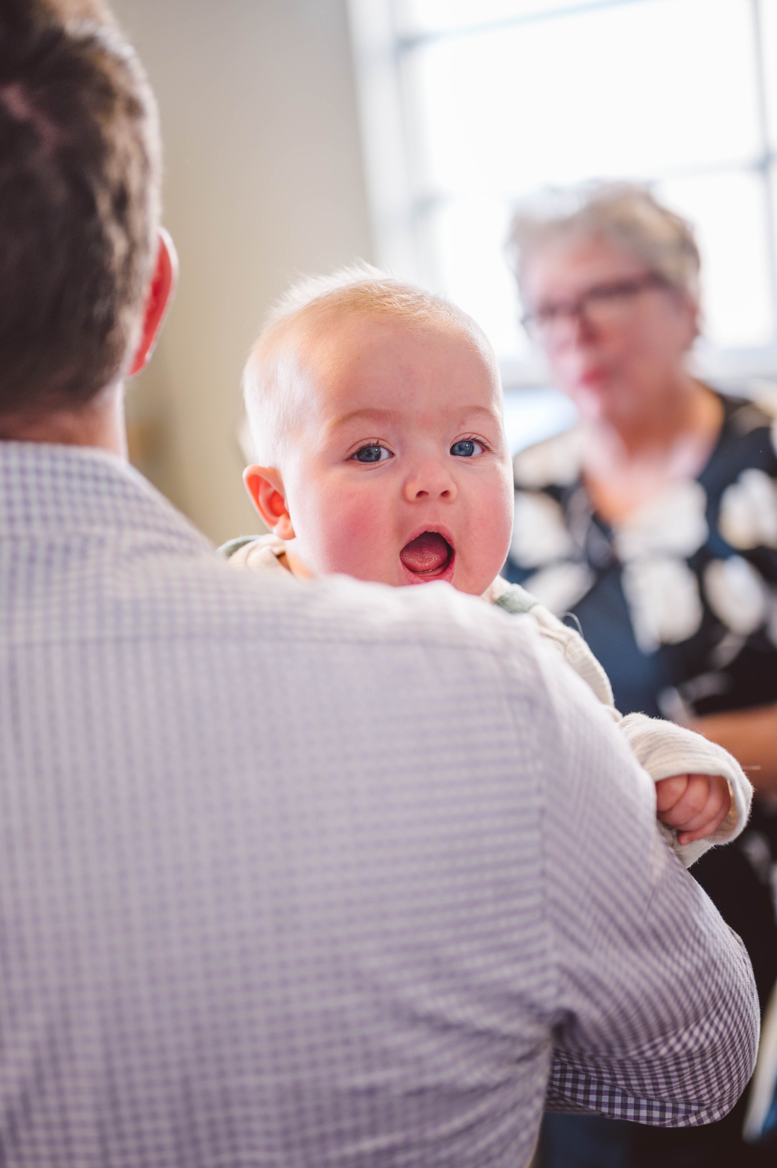 A young child with blue eyes and light hair looking surprised while being held by a man in a checkered shirt, with an older woman in a floral shirt blurred in the background.