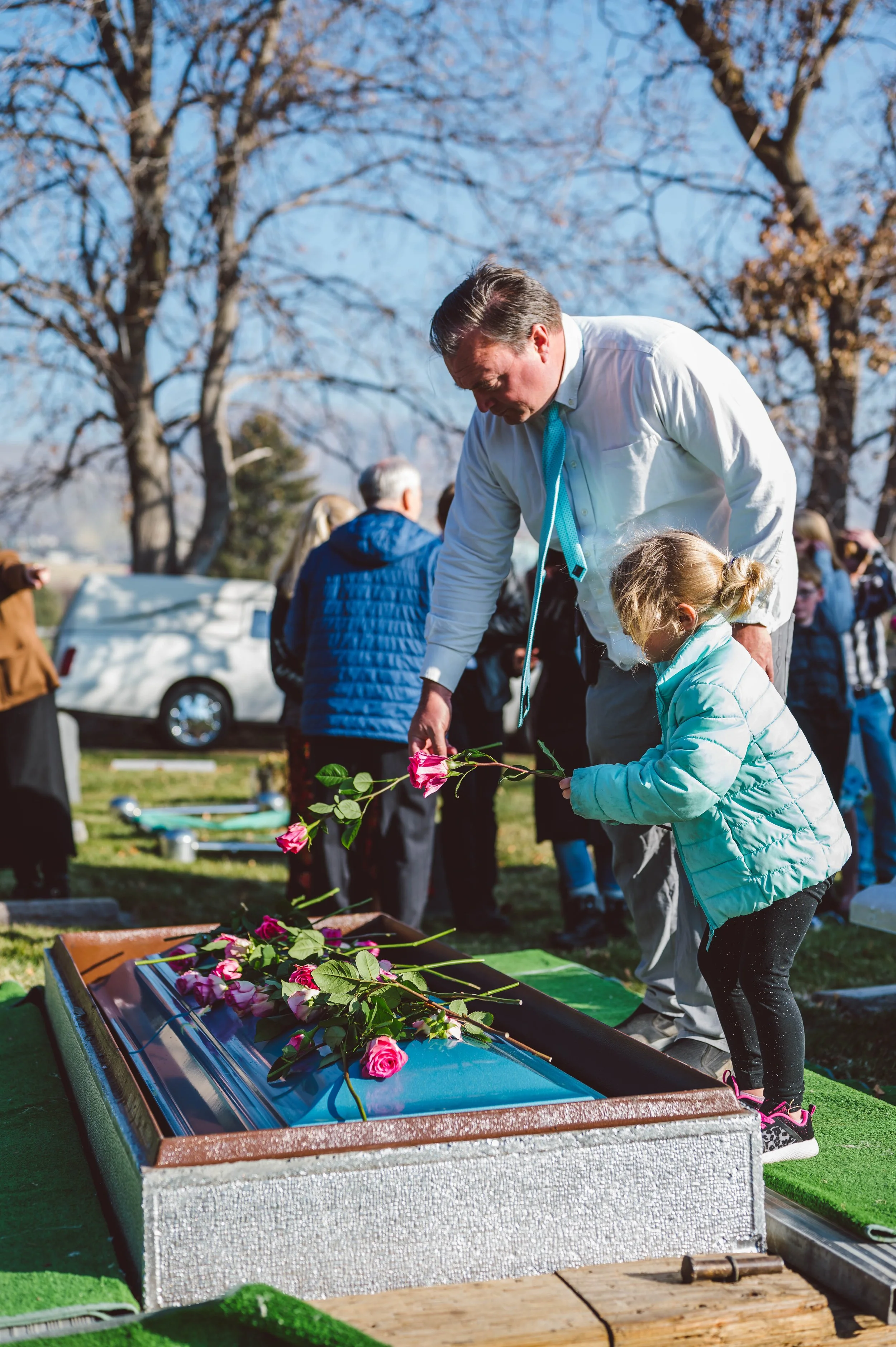 A man and a young girl place pink roses on a grave during a funeral service outdoors on a sunny day, with other people standing nearby and leafless trees in the background.