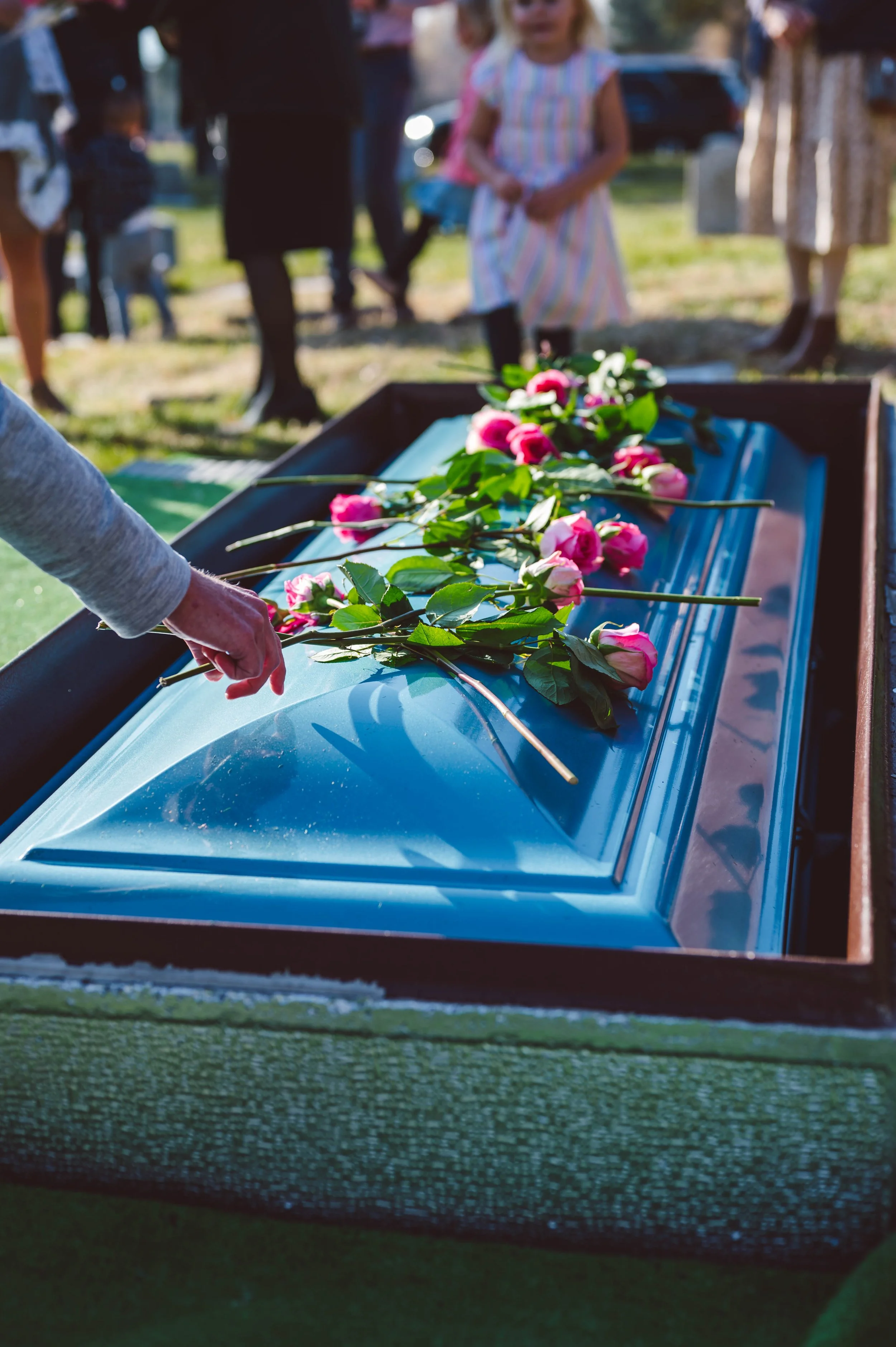 A person placing pink and white roses on a metallic surface of a large metal casket during a funeral or memorial service outdoors with grieving people in the background.