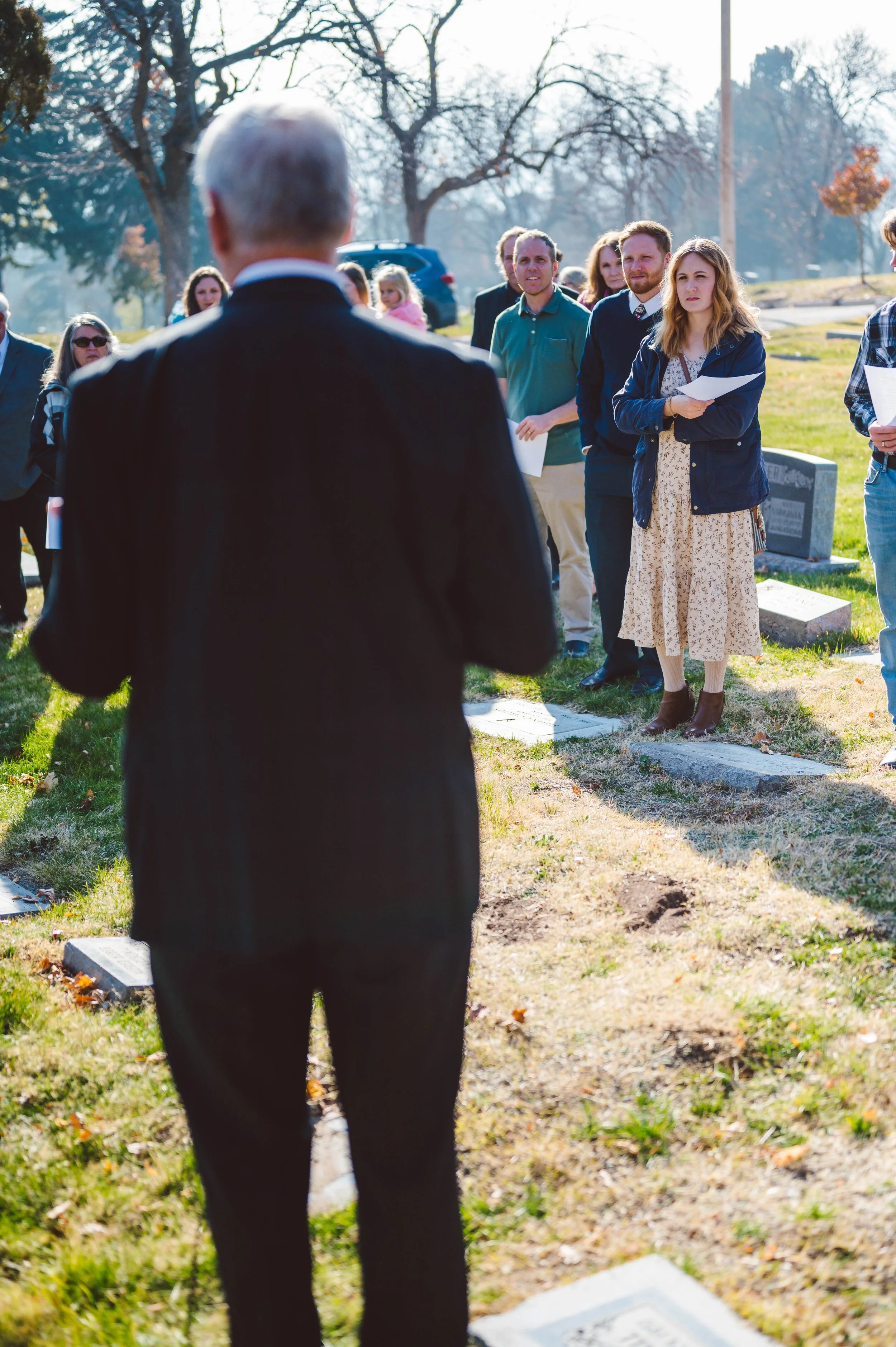People standing in a circle at a graveside funeral service, with a grave marker visible in the foreground.