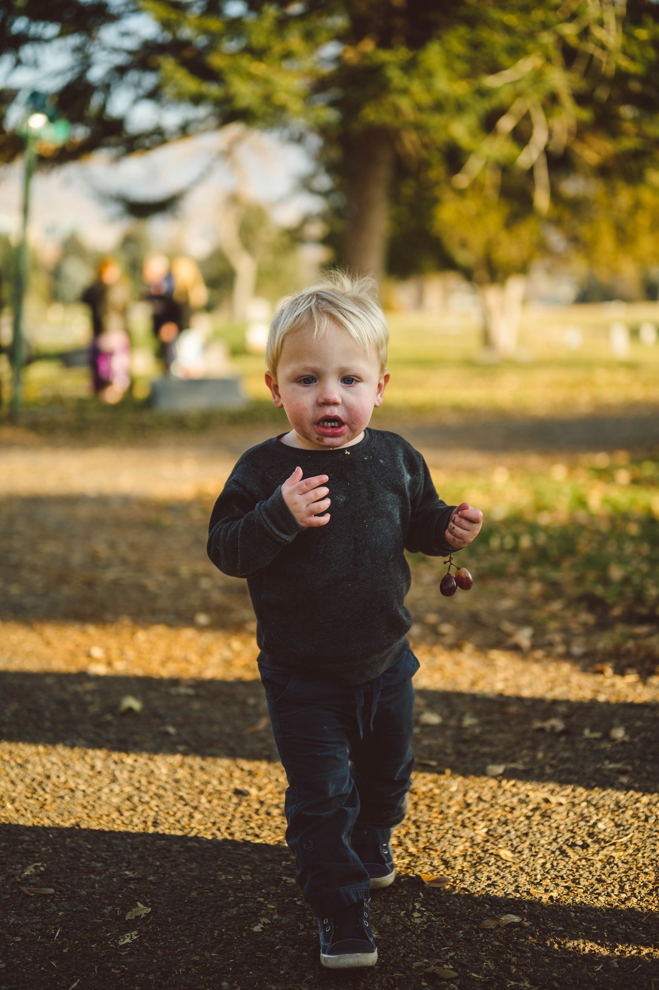 A young boy with blond hair and a black sweater running outdoors on a gravel path, holding a small branch with a berry, with trees and people in the background.