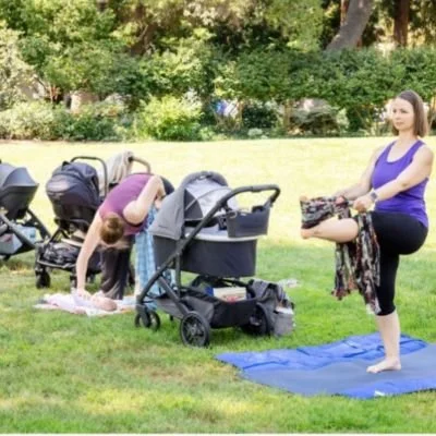 Some moms outside on the grass, with baby strollers next to them while doing yoga.