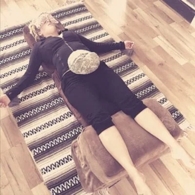 A woman on a rug at home, relaxed lying on her back, with feet elevated and heat pack on her stomach relaxing with arms out to her sides.