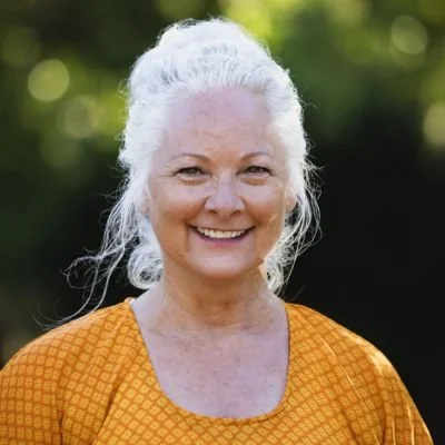 Martie headshot smiling at camera, with white hair up behind her, and yellow and orange dress shirt.