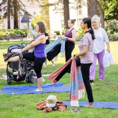 A few parents outside on the grass holding yoga straps, with baby strollers next to them while doing yoga.