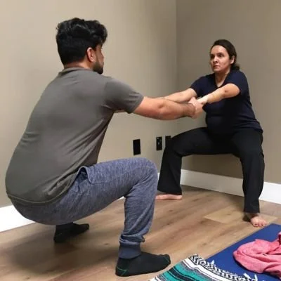 A couple in a deep squat facing each other, holding each others hands with arms out in front of them to help balance.