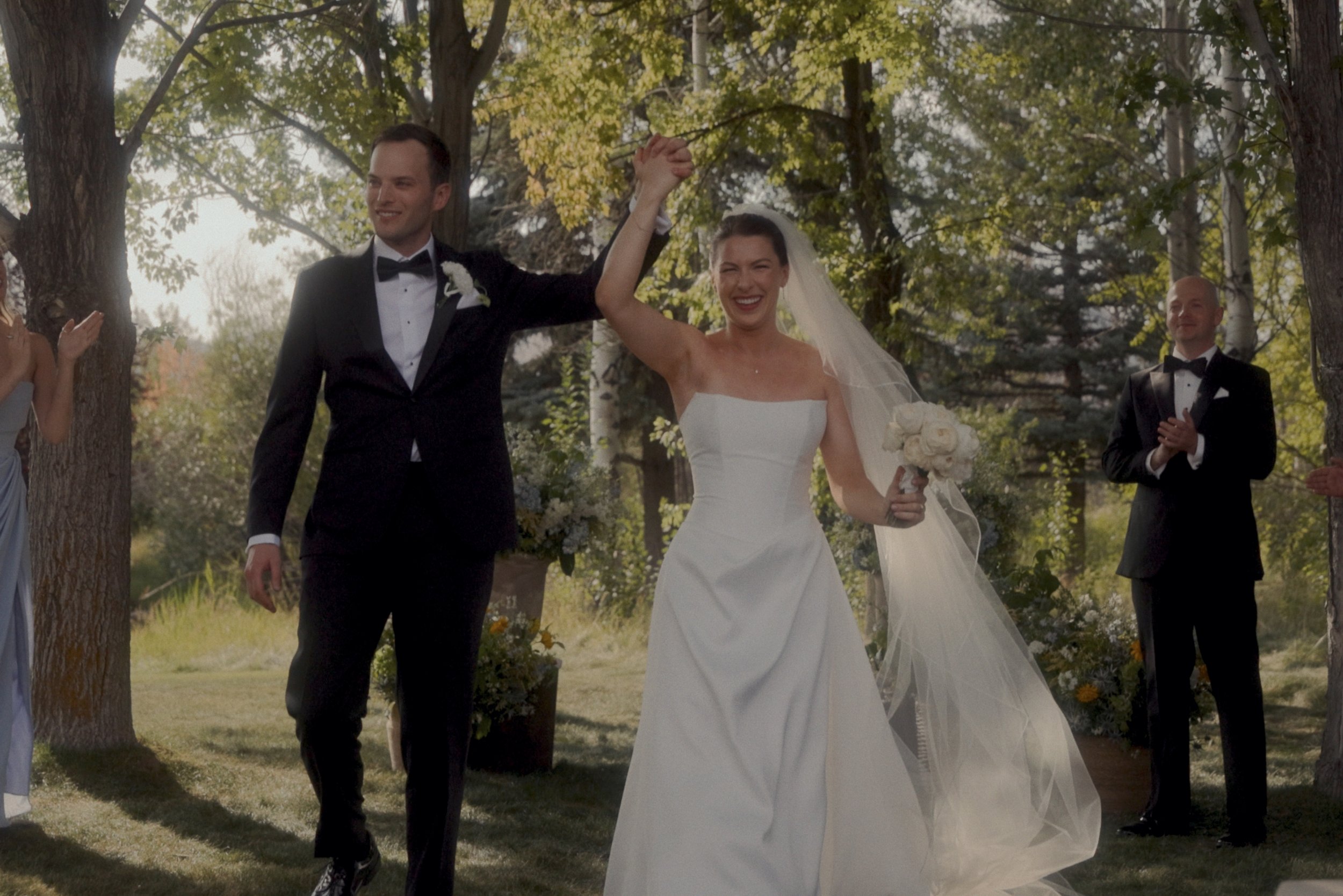 Newlywed bride and groom celebrating outdoors under trees with wedding guests applauding