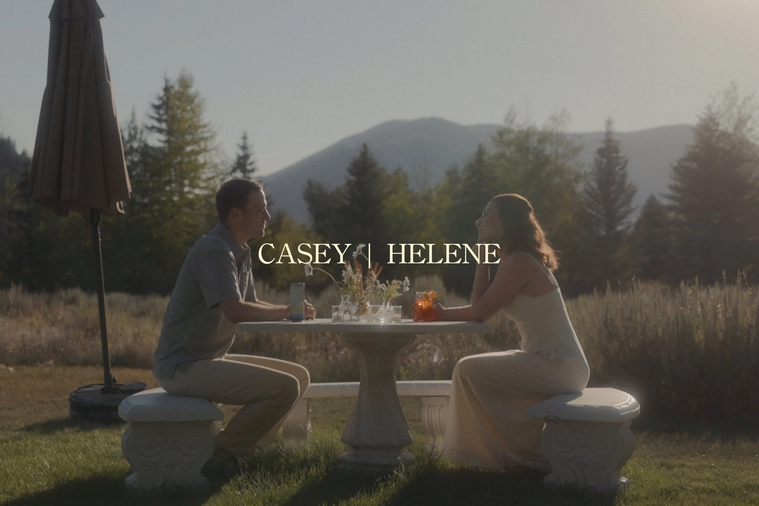 Couple dining at a picnic table in a mountain meadow at sunset, with an umbrella and floral centerpiece; text reads “Casey | Helene.”