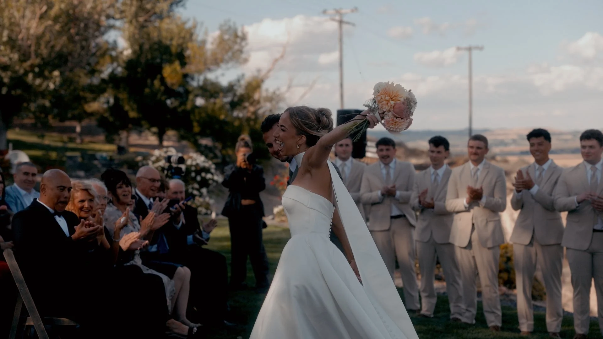 Bride holding a bouquet high celebrating after a wedding ceremony surrounded by applauding guests in an outdoor setting.