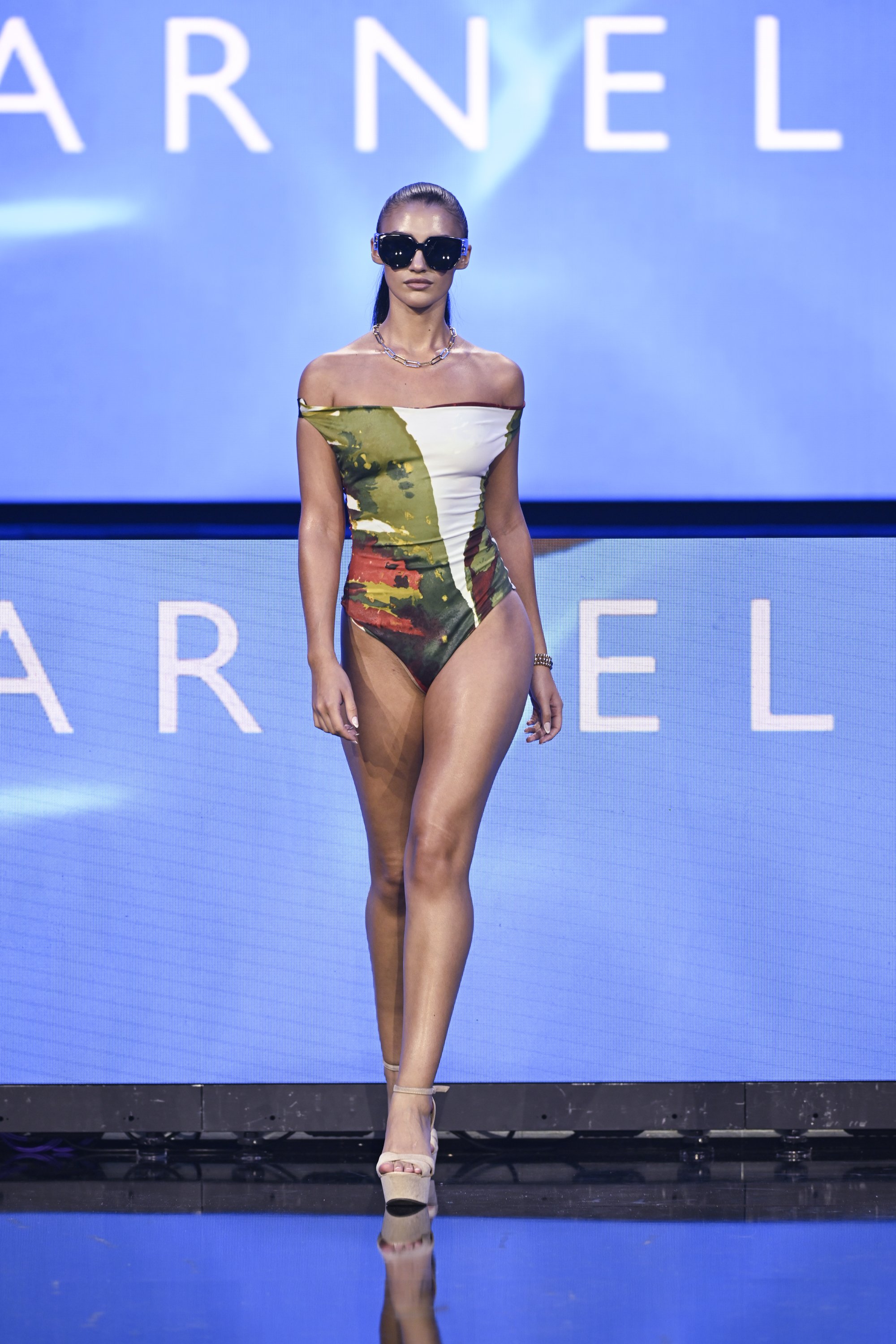 A female model walking on a runway in a camouflage and white swimsuit, wearing large black sunglasses, a necklace, and white high heels, with a blue background displaying partially visible white text.