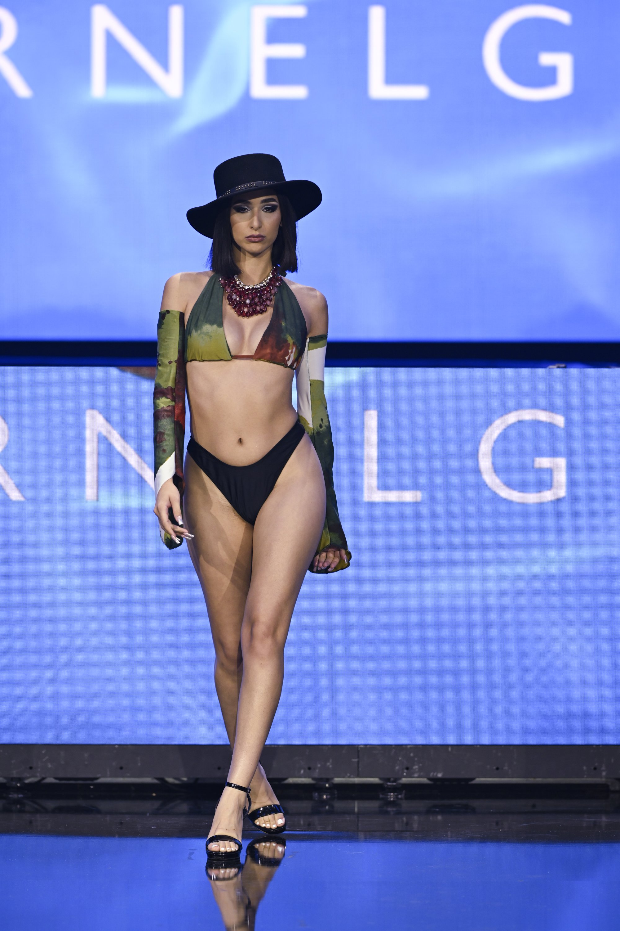A female model walking on a runway wearing a bikini with a wide-brimmed hat, a large red necklace, and patterned long sleeves, with a blue backdrop displaying the word "RNELG" interrupted by the model.