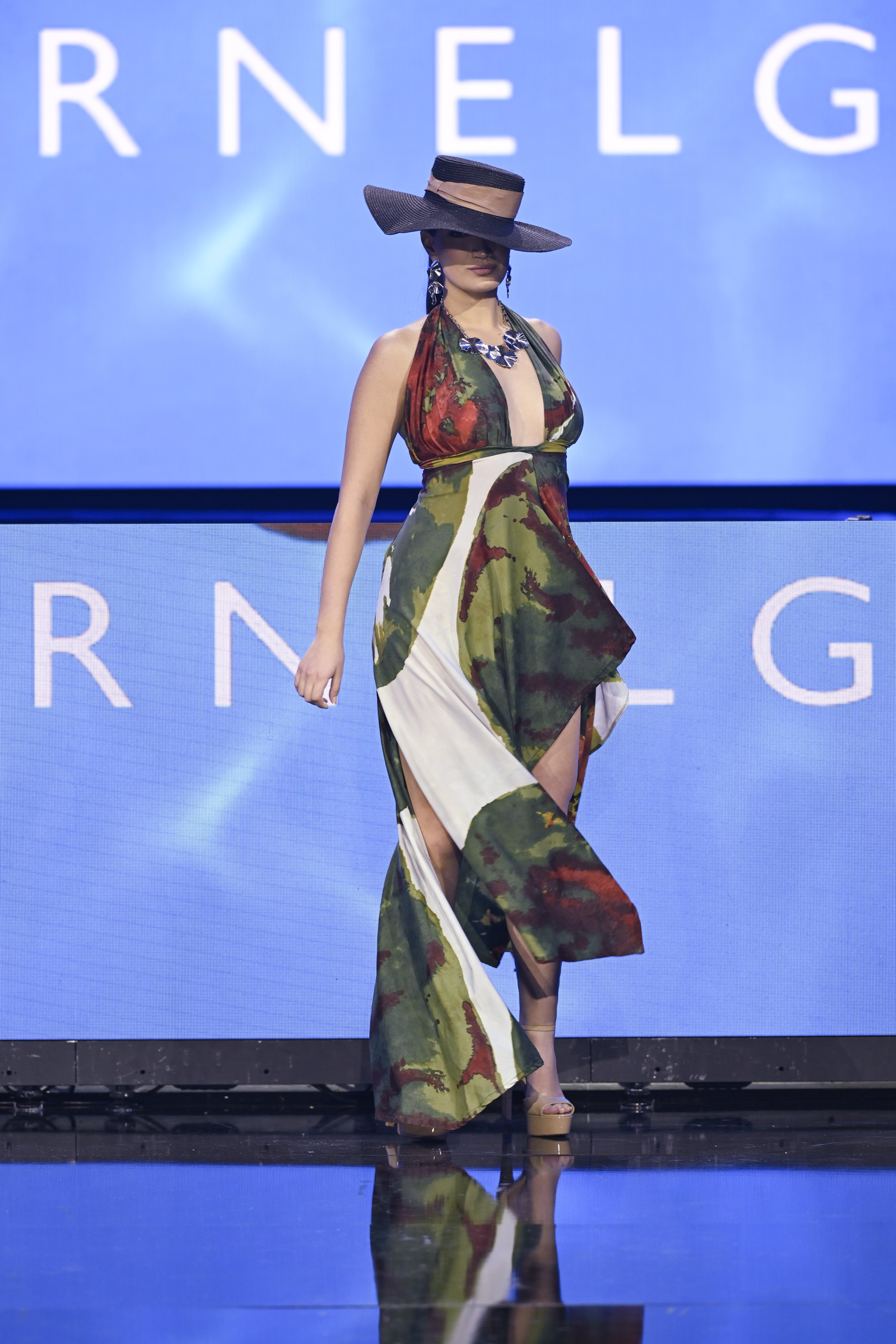 A woman walking on a runway wearing a large brimmed hat, a colorful halter dress with a slit showing her leg, and high heels. The background features a blue screen with partial white text.