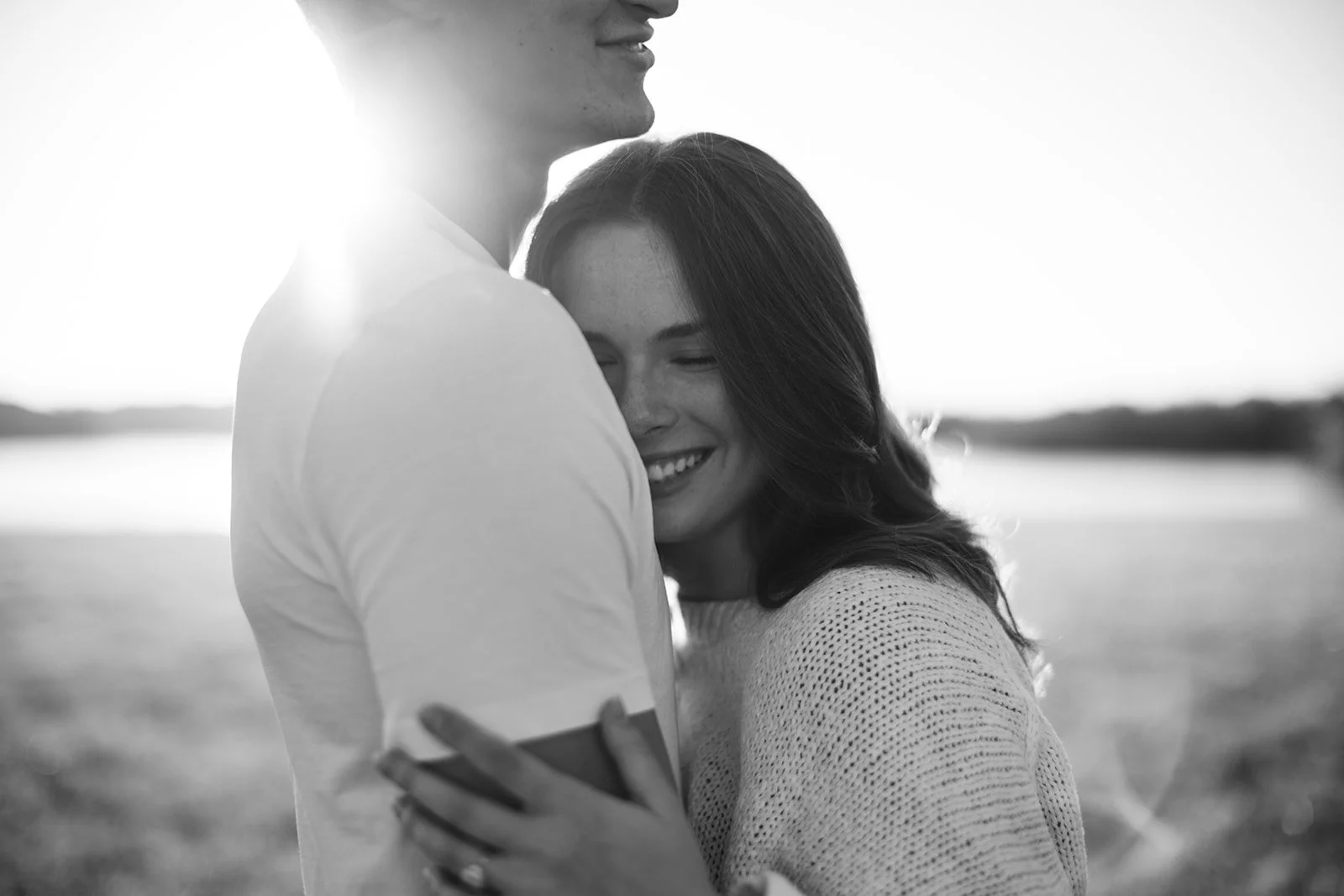 A black and white photo of a couple hugging outdoors, with the woman smiling and the man partially visible from the nose down, the sun shining in the background.